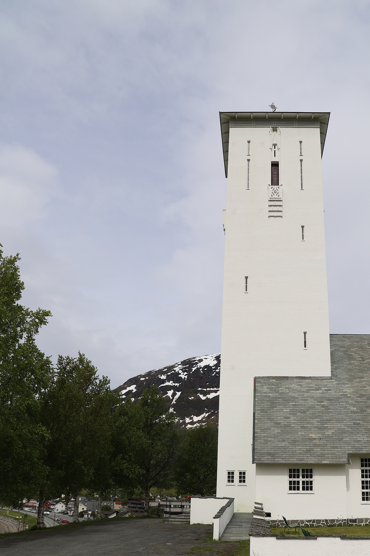Et høyt, hvitt kirketårn med smale vinduer står ved siden av grønne trær og et snødekt fjell i bakgrunnen under en overskyet himmel.