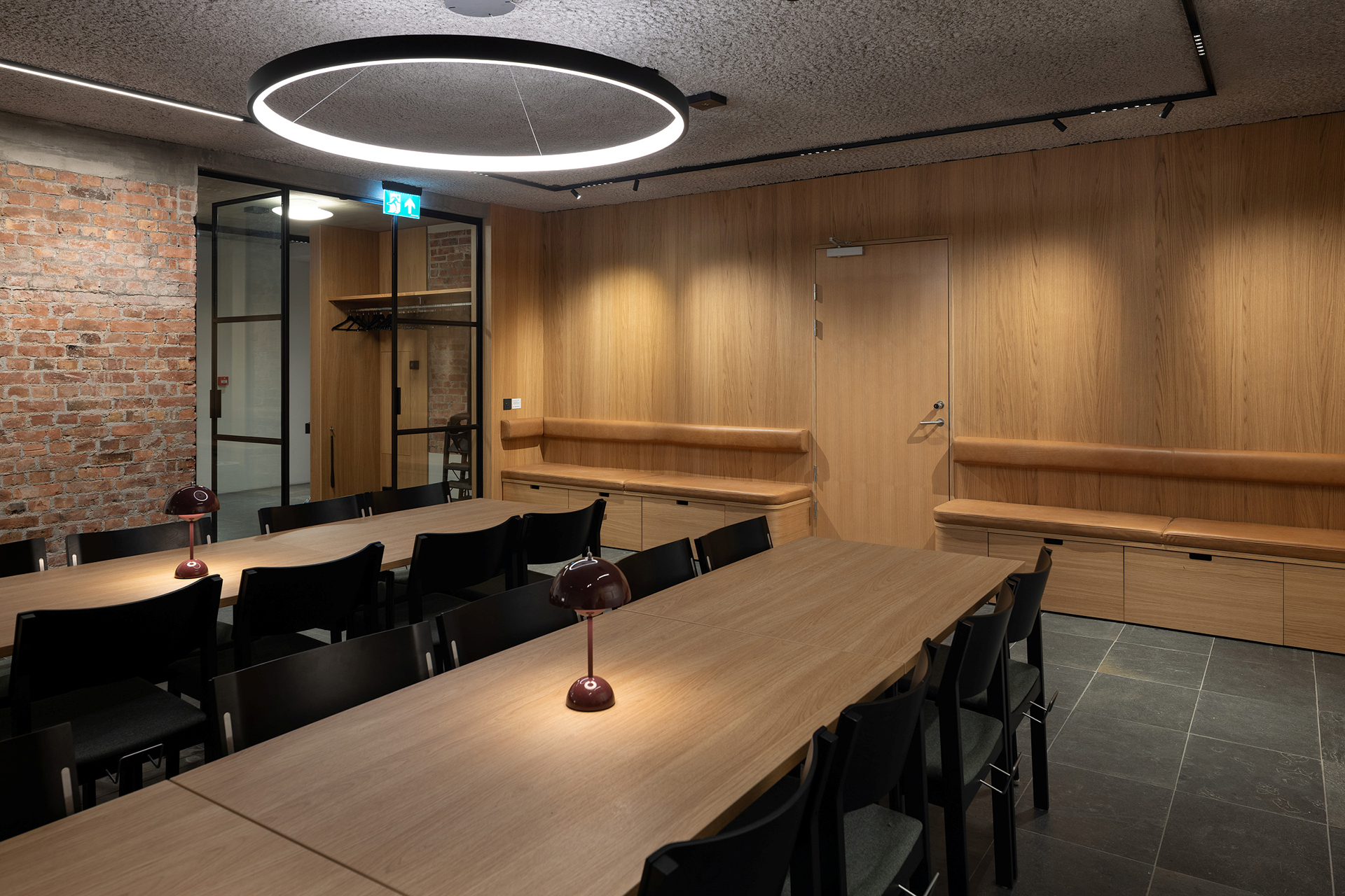 Moderne konferanserom med trepanel på veggene, teglstein i aksenten, svarte stoler rundt et langbord, innebygde sittebenker og en sirkelformet taklampe.