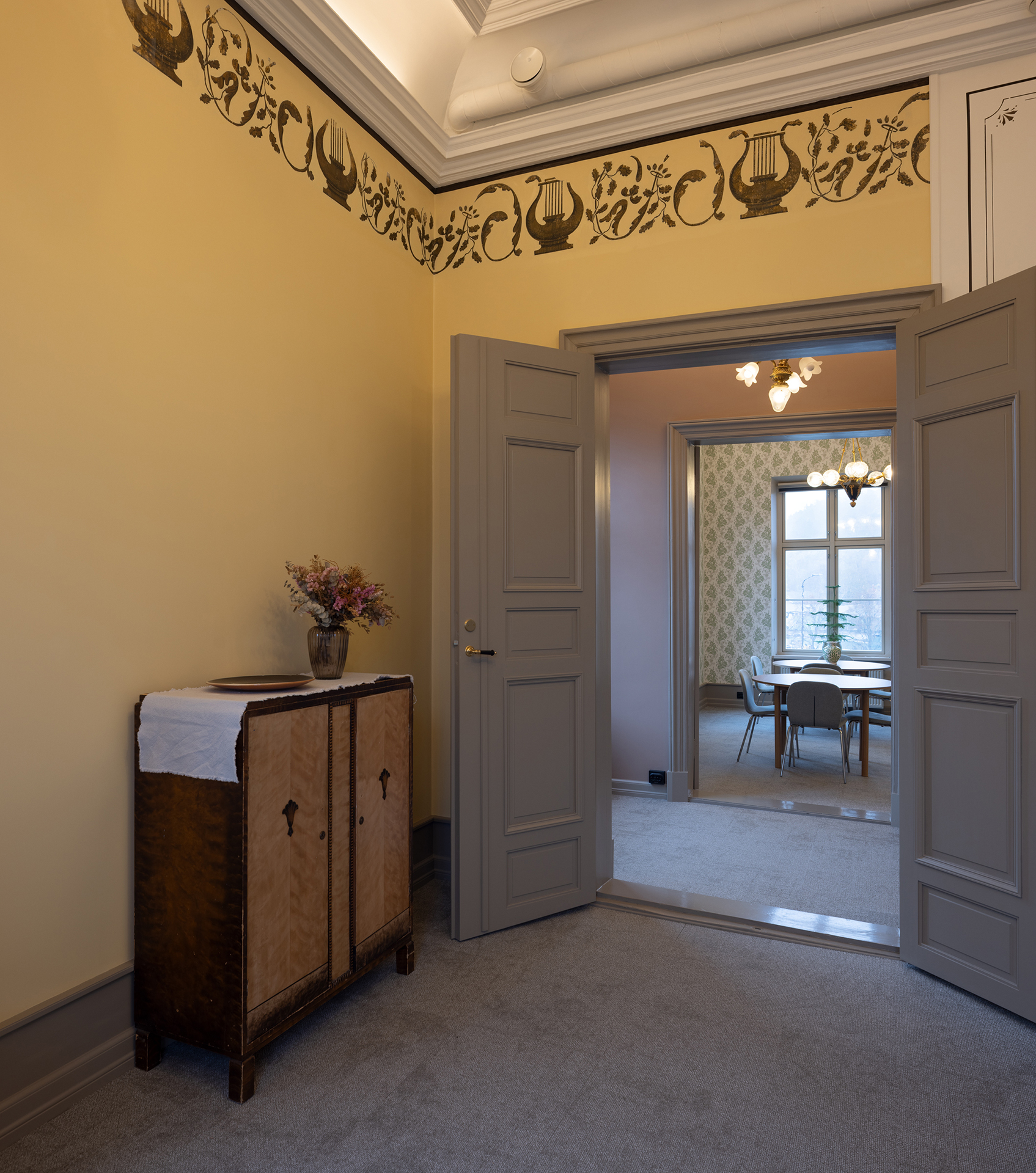 En gang med lysegule vegger, dekorativ bård, et treskap med en vase med blomster og doble dører som leder inn til et rom med bord og stoler.