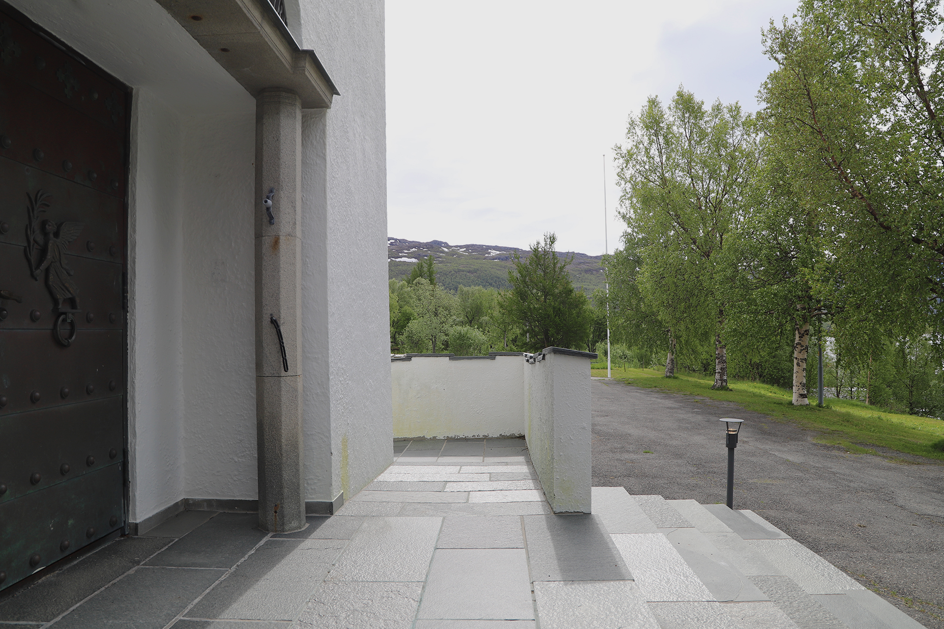 Utsikt fra en steintrapp utenfor en hvit bygning, med trær, gress og en asfaltert sti; i bakgrunnen skimtes fjell under en overskyet himmel.