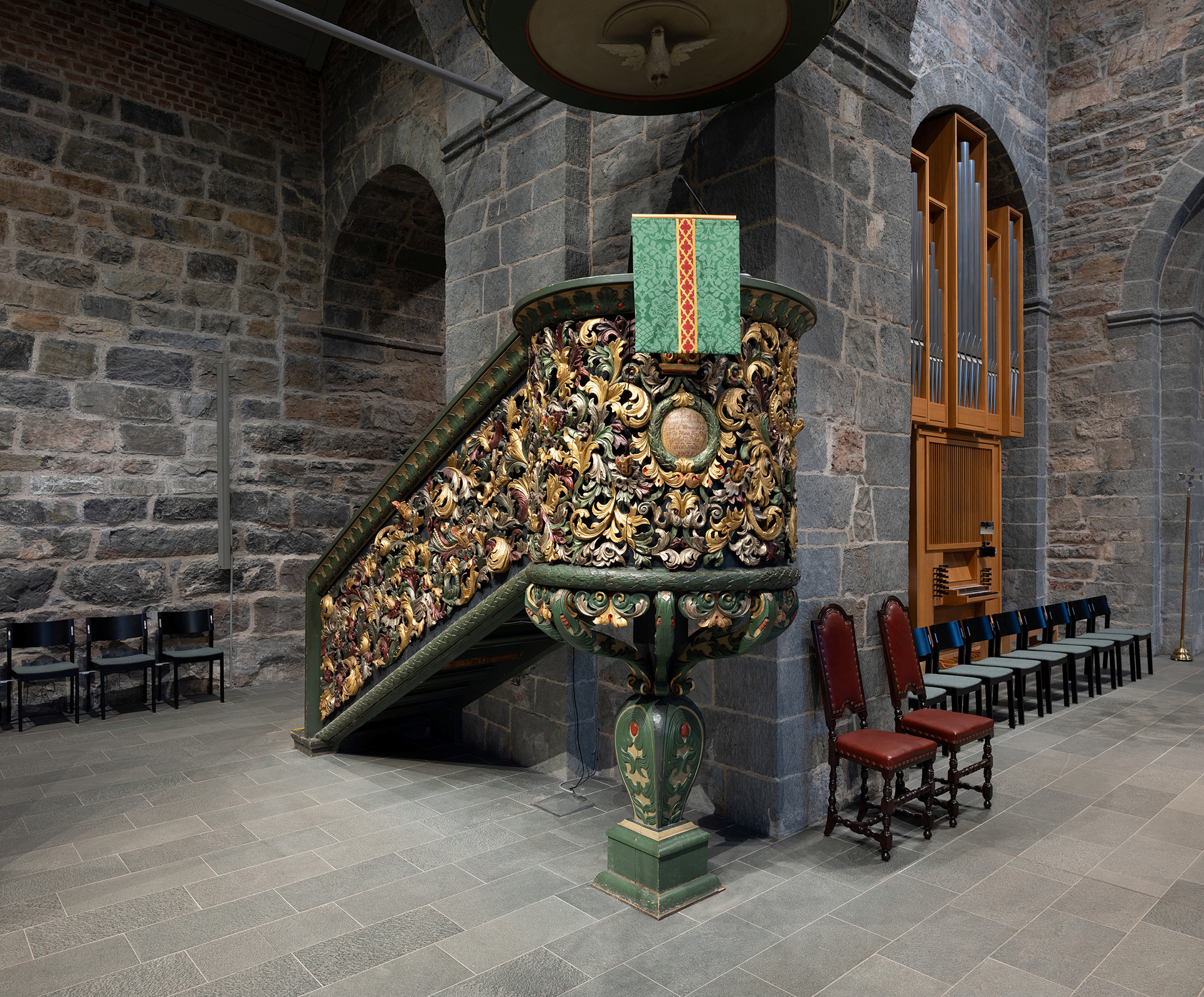 Utsmykket, dekorativ prekestol i tre med intrikate utskjæringer og et grønt klede, plassert i et kirkerom av stein i nærheten av stolrader og orgel.