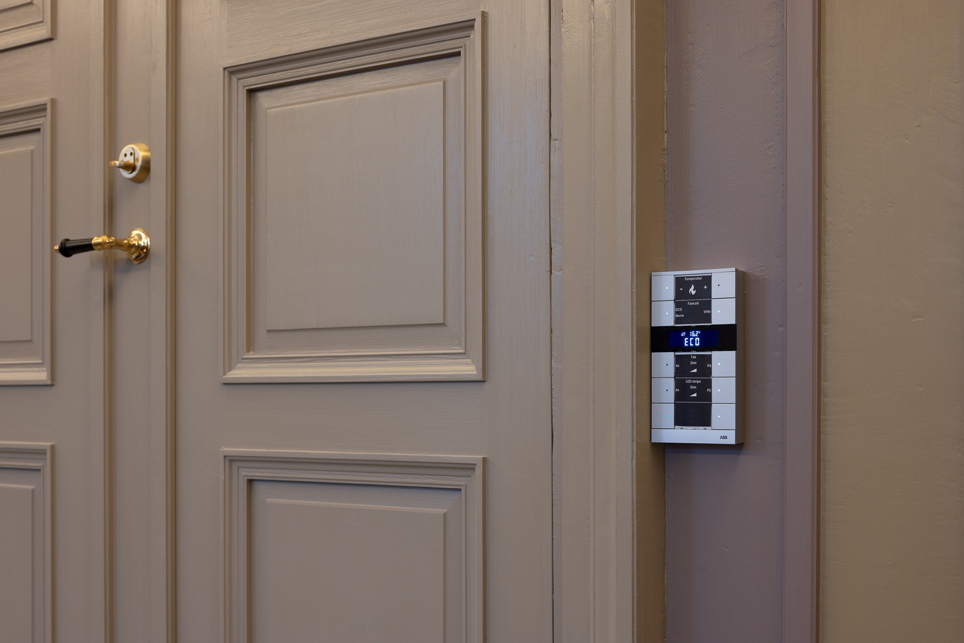 En lukket brun dør med gullbeslag er plassert ved siden av et veggmontert elektronisk kontrollpanel som viser klokkeslettet.