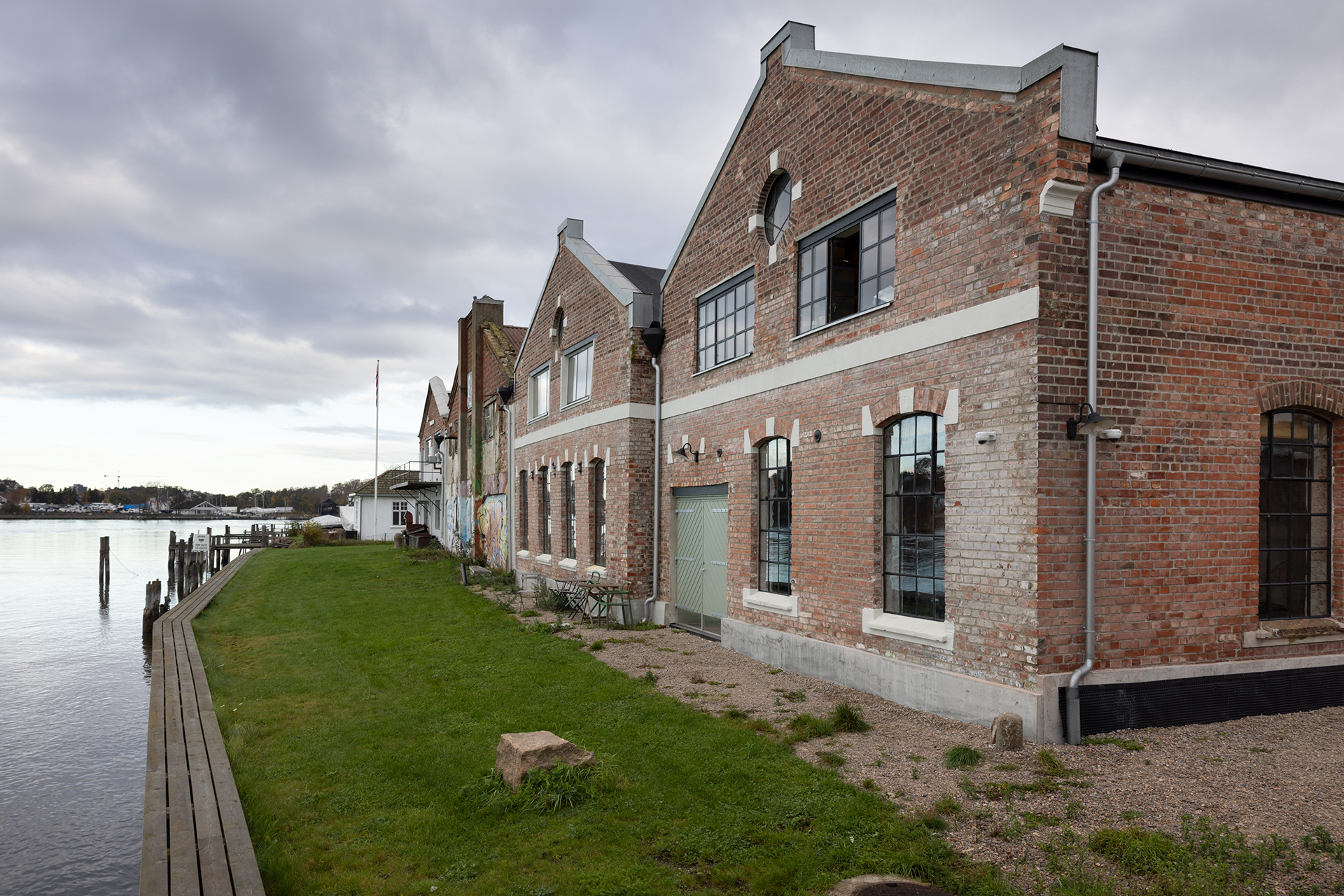 En industribygning i teglstein med store vinduer ligger ved siden av et gressområde og en trebrygge ved vannet under en overskyet himmel.
