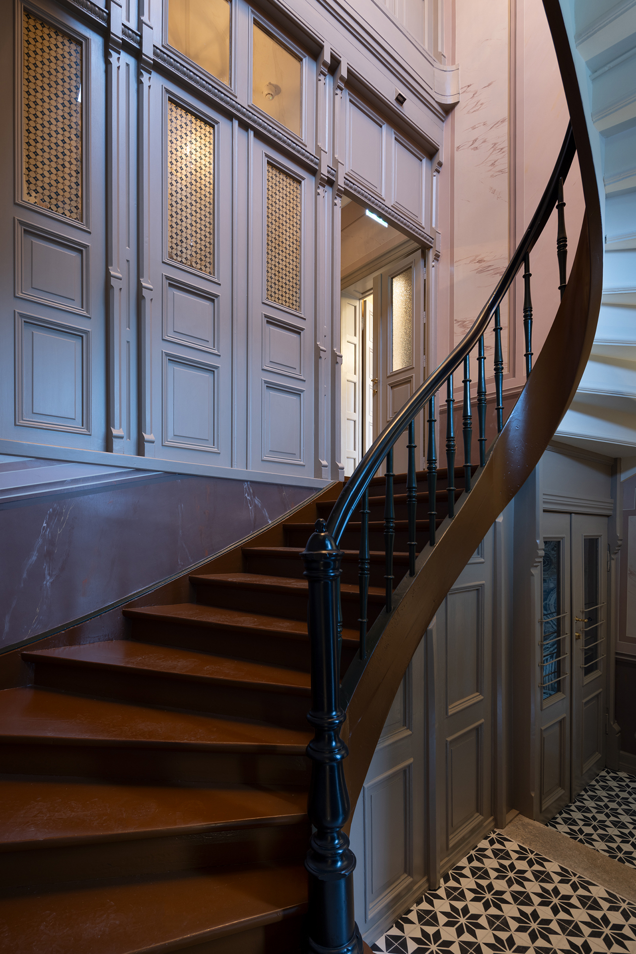 Buet tretrapp med svart rekkverk i en gang med panelvegger og geometrisk mønstrede gulvfliser.