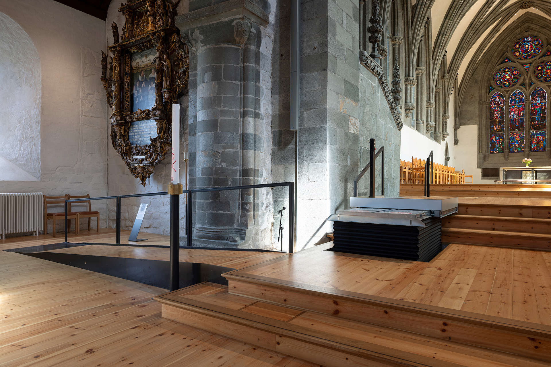 Moderne tilgjengelegheit i mellomalderkyrkje 12 Interiøret i en historisk kirke med steinsøyler, tregulv med trappetrinn, en moderne rullestolheis, glassmalerier og utsmykket veggkunst.