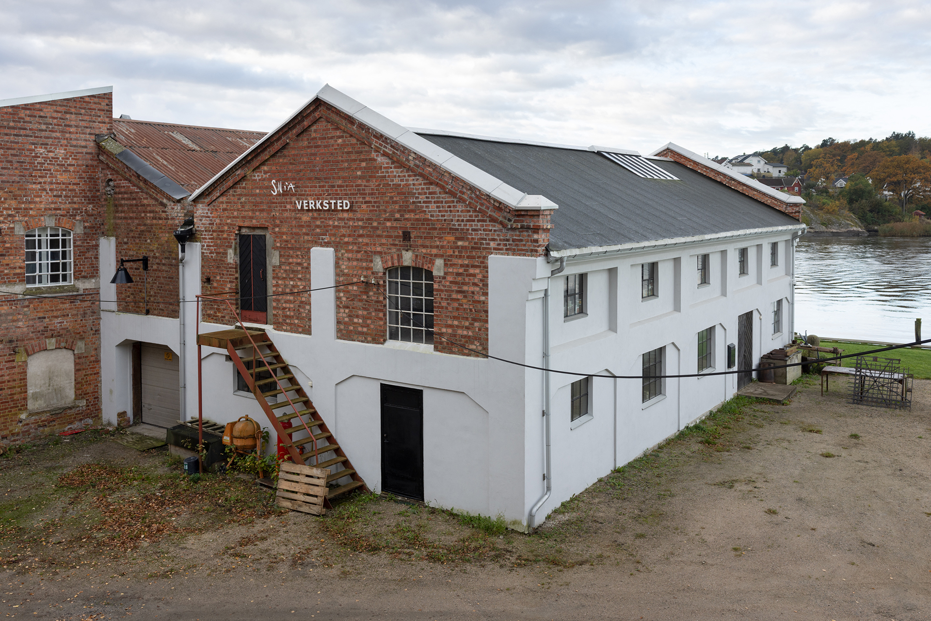 Sirkulærøkonomi i praksis 17 En toetasjes bygning i murstein og hvitt med navnet Verksted står ved en elv, med en utendørs trapp som fører til en dør i andre etasje og en liten terrasse til høyre.