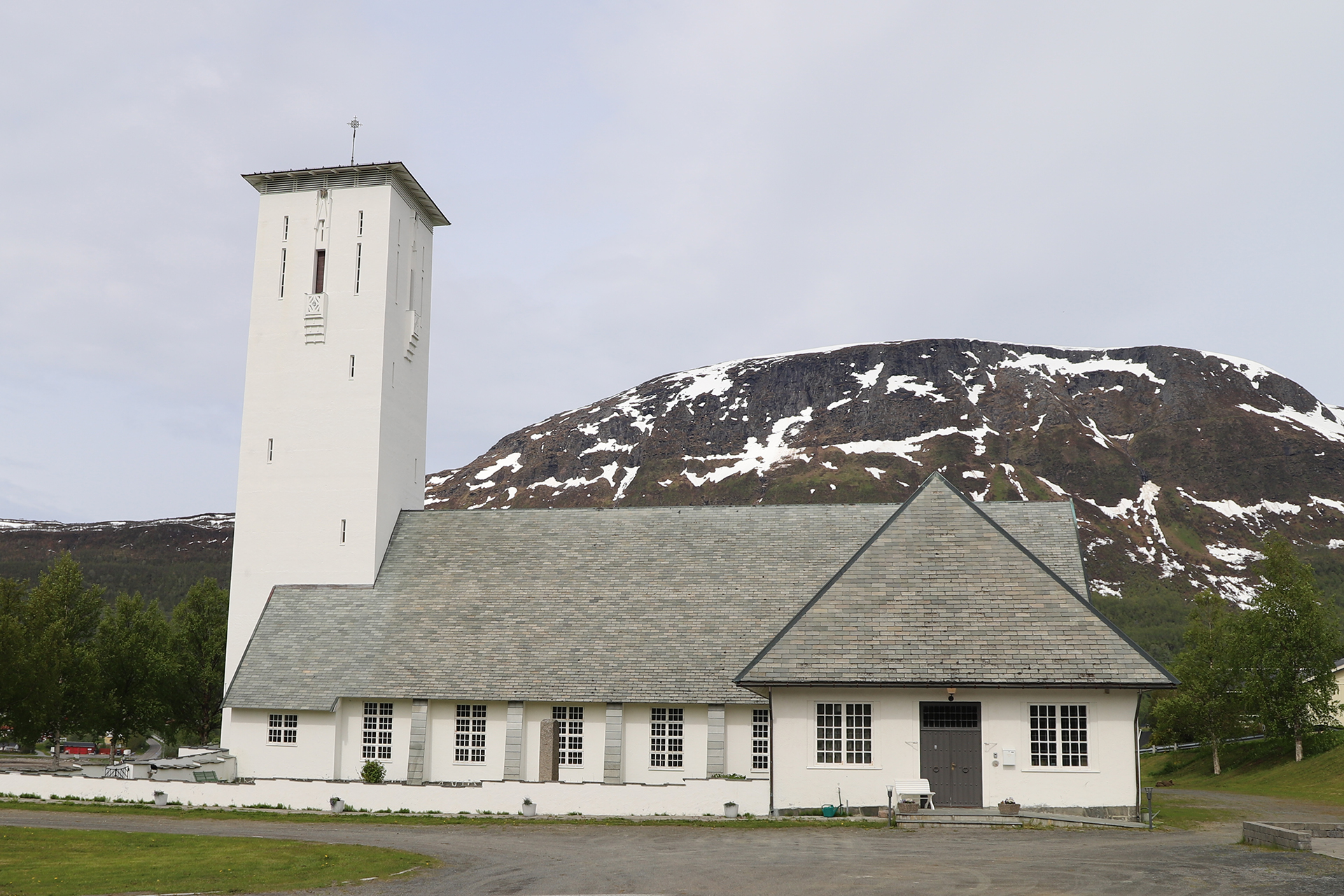 En hvit kirke med et høyt rektangulært tårn står foran et snødekt fjell, omgitt av grøntområder og en overskyet himmel.