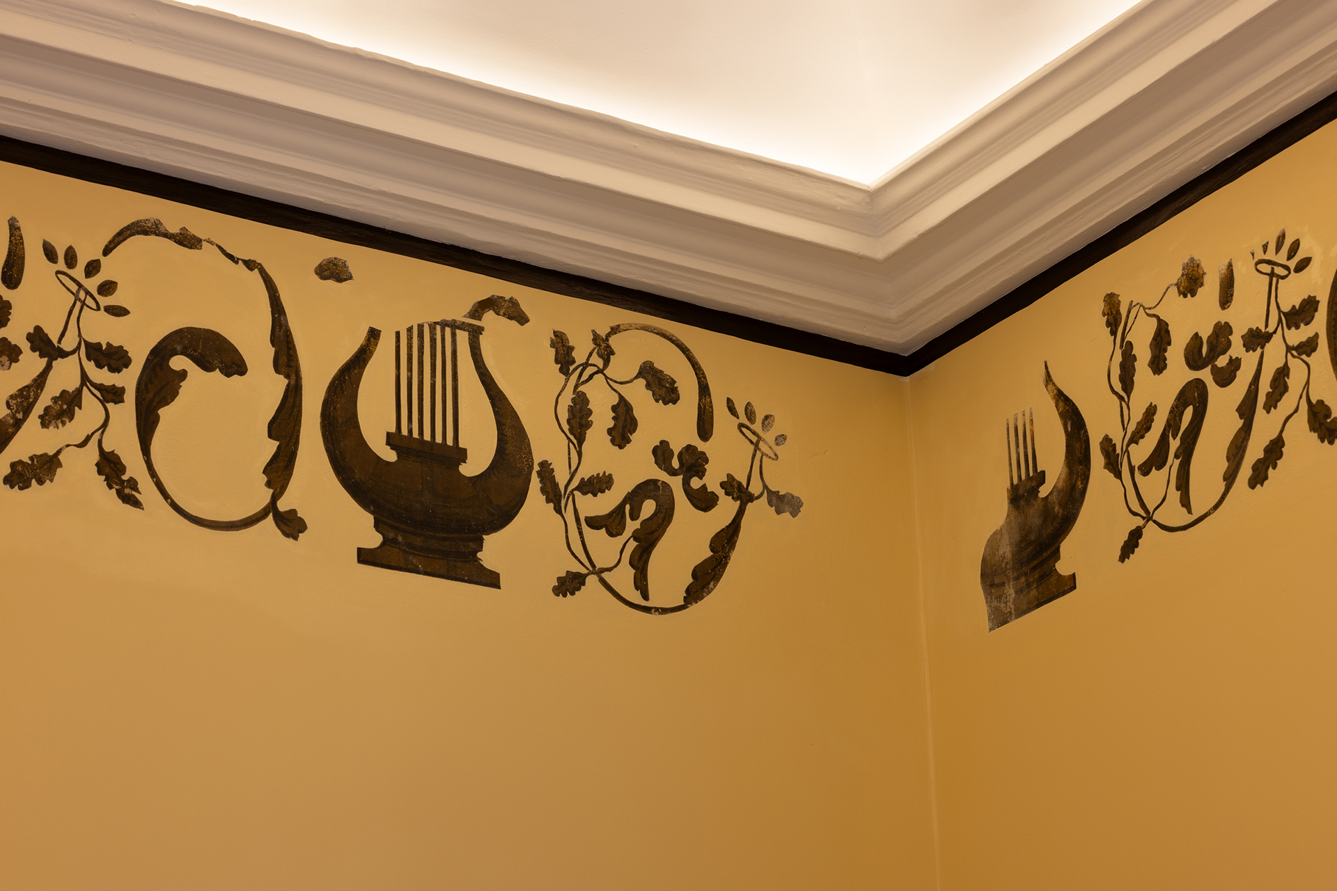 Utsmykkede svarte sjablongmotiver, inkludert lyrer og blomstermotiver, dekorerer det øvre hjørnet av en gul vegg nær et hvitt tak med kronlist.