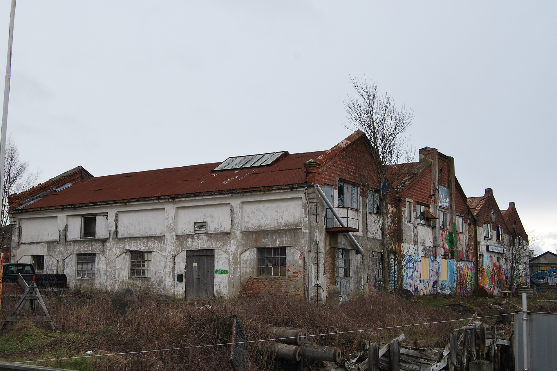 En rekke gamle, nedslitte bygninger med avskallet maling, rustne tak og graffiti på veggene, omgitt av overgrodd vegetasjon under en overskyet himmel.