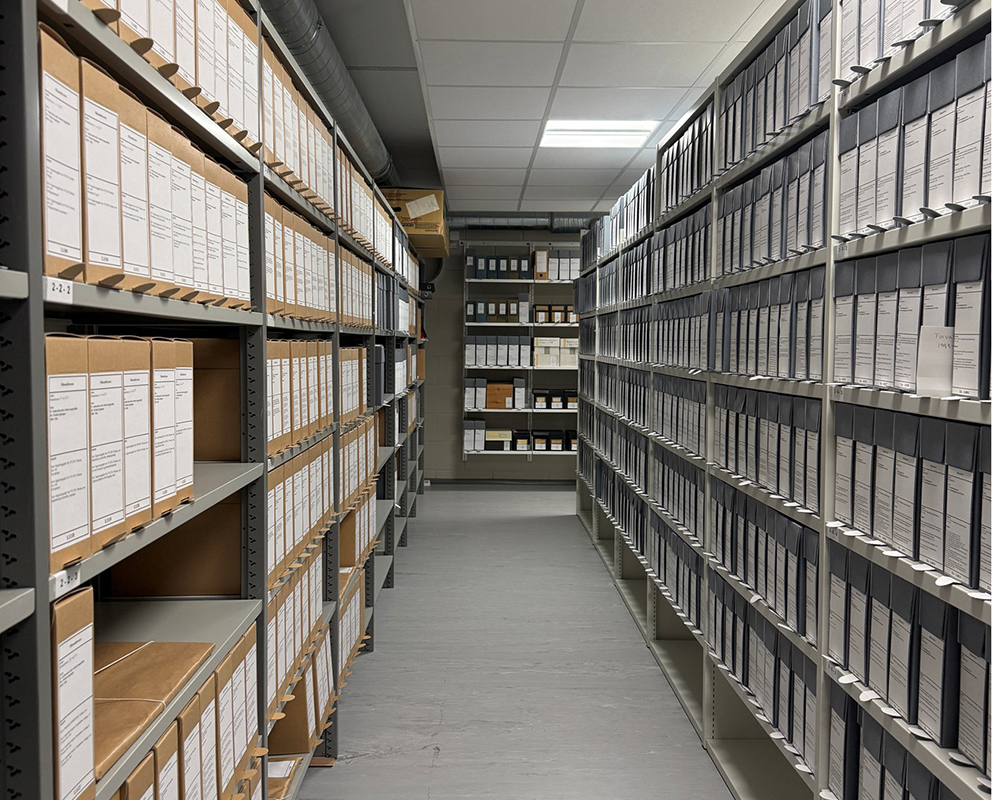Et lagerrom med hyller fulle av merkede arkivbokser og mapper på rekke og rad langs veggene.