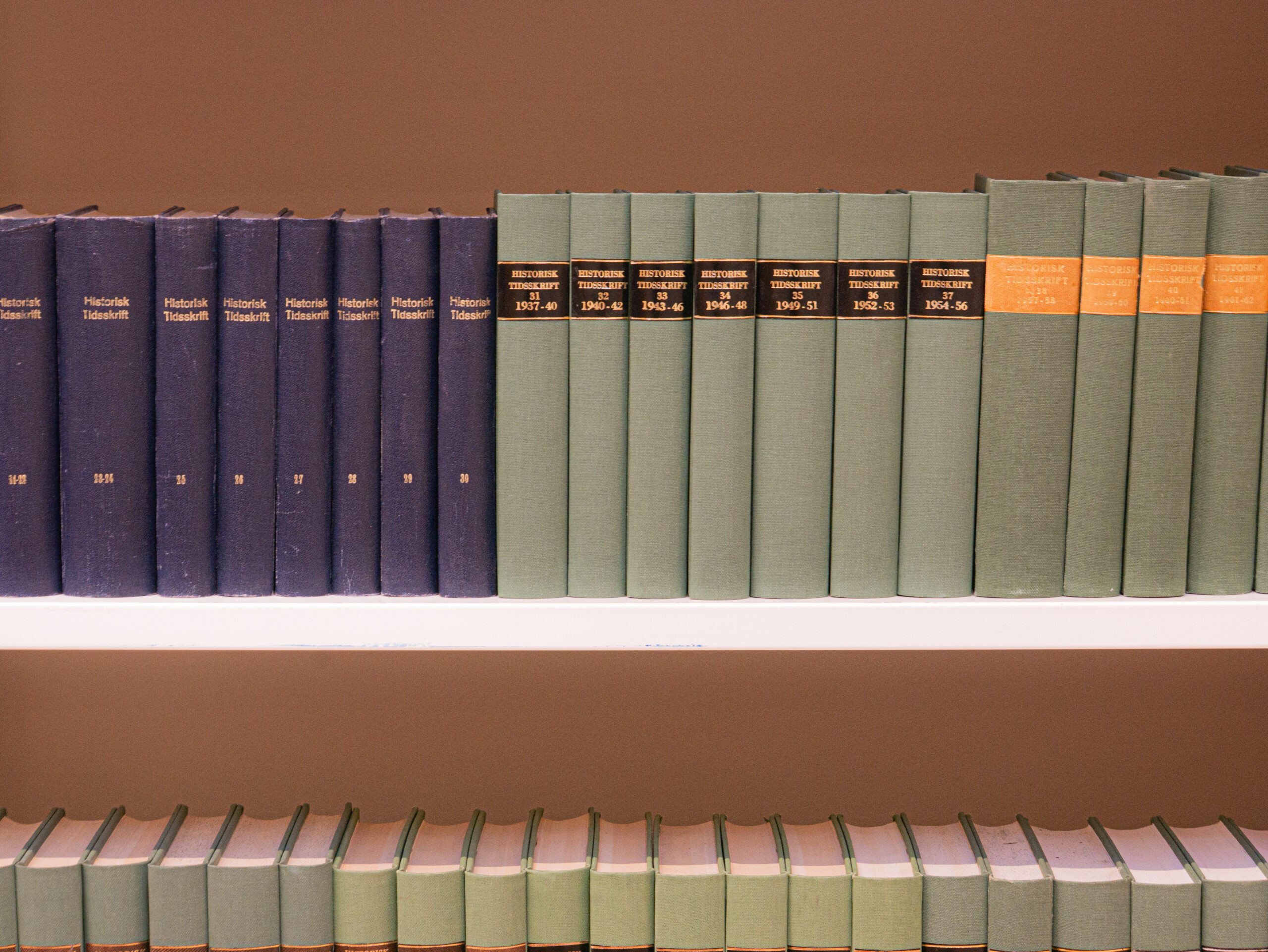 En rekke blå og grønne innbundne bøker med gullfargede og svarte etiketter står sirlig plassert på hvite hyller.