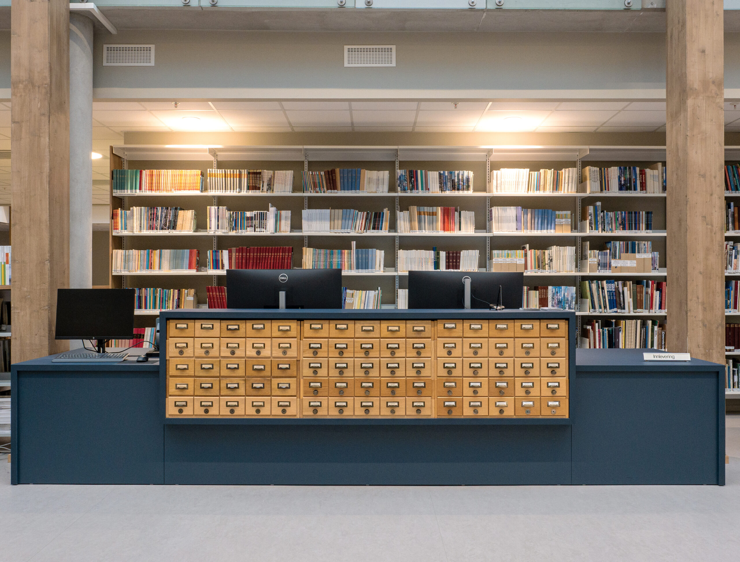 En biblioteksdisk med to dataskjermer, et gammelt kortkatalogskap og bokhyller fylt med bøker i bakgrunnen.