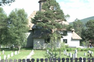 Dombås kyrkje står bak trær og ein liten kyrkjegård med kvite gravsteinar og eit mørkt stakittgjerde i forgrunnen.