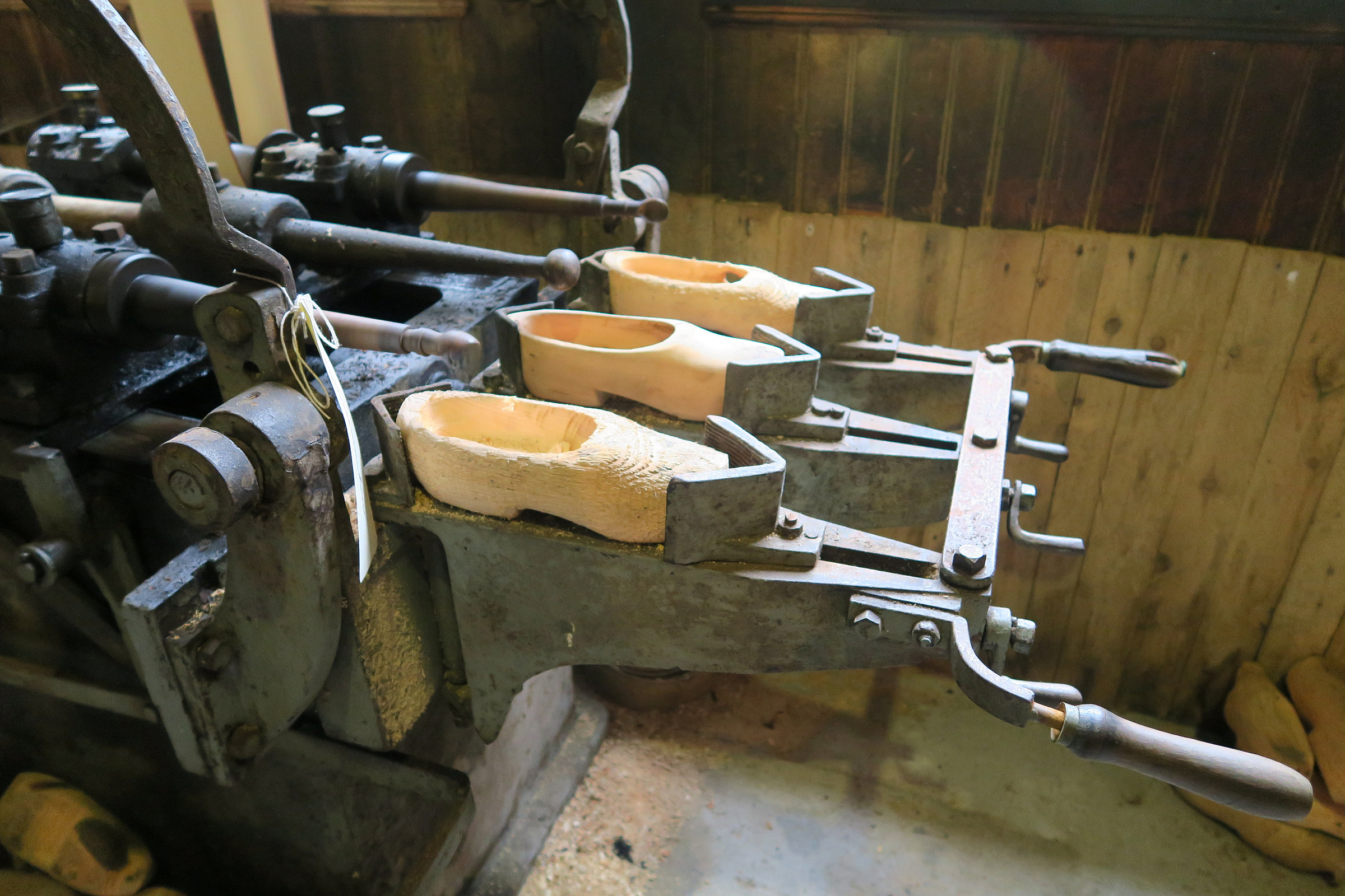 Nærbilde av en gammel maskin som holder tre uferdige tresko i metallklemmer, plassert i et verksted.