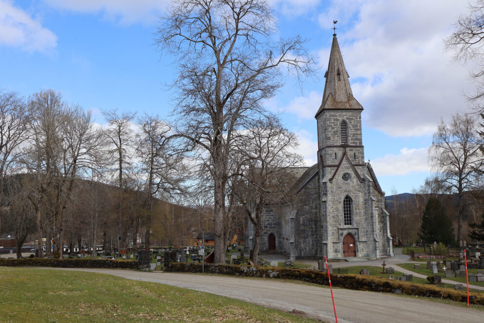 I disse kirkene vil du merke forskjell til jul 6 En steinkirke med et høyt kirketårn står blant løvløse trær, med en kirkegård og en buet grussti i forgrunnen under en delvis overskyet himmel.