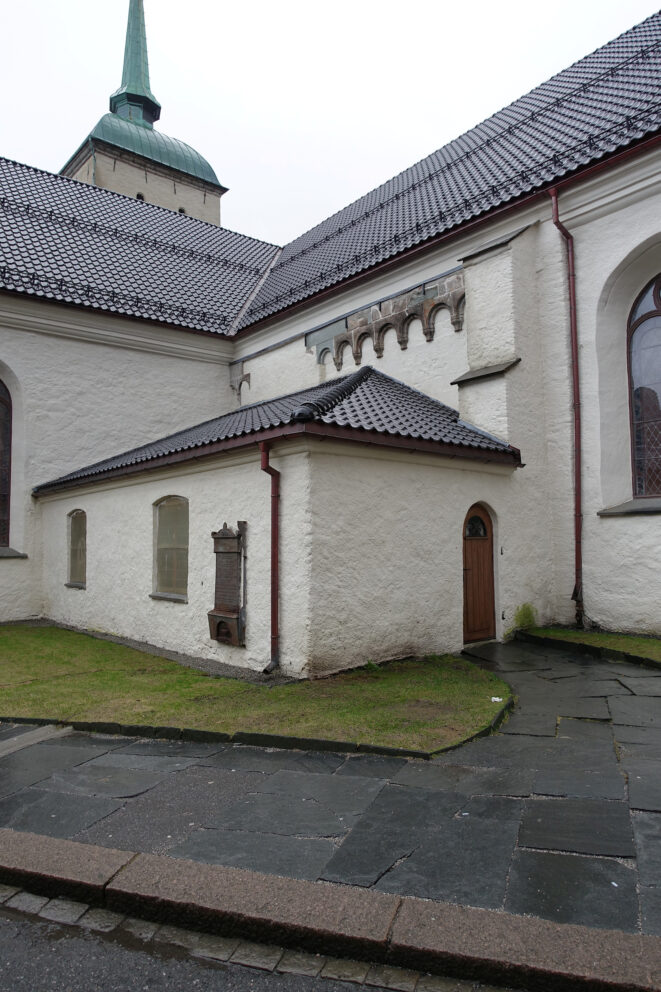 I disse kirkene vil du merke forskjell til jul 9 En hvit steinbygning med bratt, mørkt tak, buede vinduer, en tredør og en metallplate på veggen, med vått fortau og gress foran.