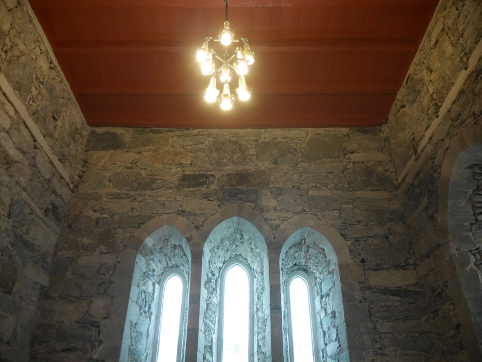 I disse kirkene vil du merke forskjell til jul 10 Tre høye, smale, buede vinduer i en steinvegg under et rødt tretak, opplyst av en hengende lysarmatur.
