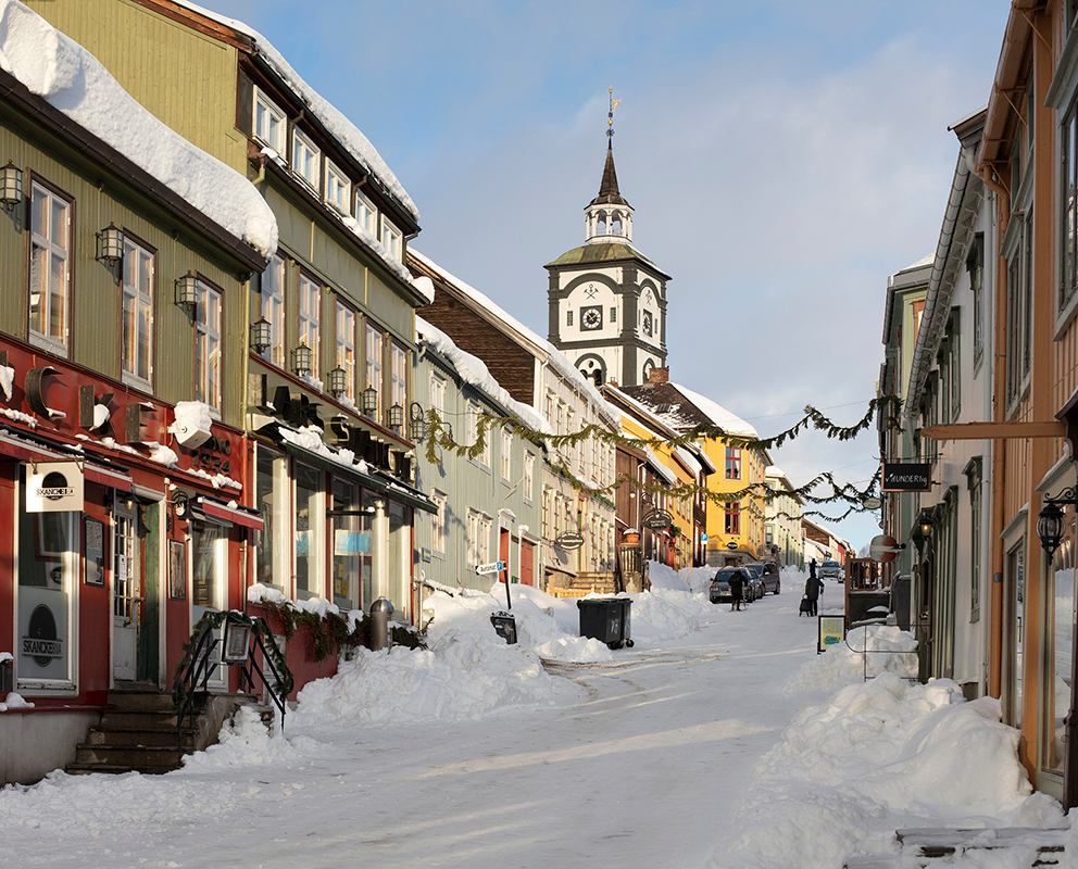Snødekte gater med fargerike trebygninger fører til en kirke med klokketårn under en delvis overskyet himmel.