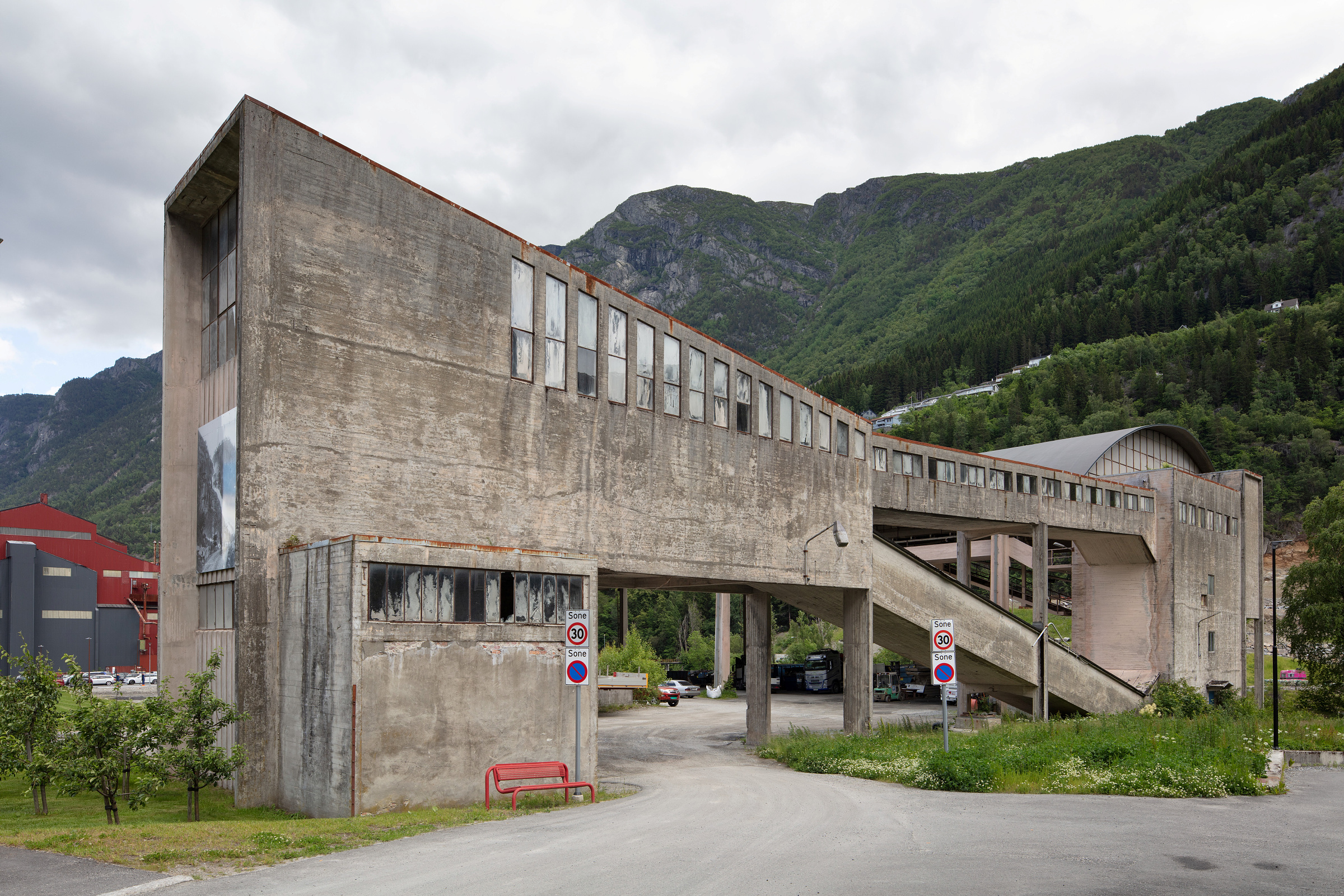 Gangbro i betong med et kantete, industrielt design, opphøyd av søyler, i et fjellområde med skyet himmel og grøntområder.
