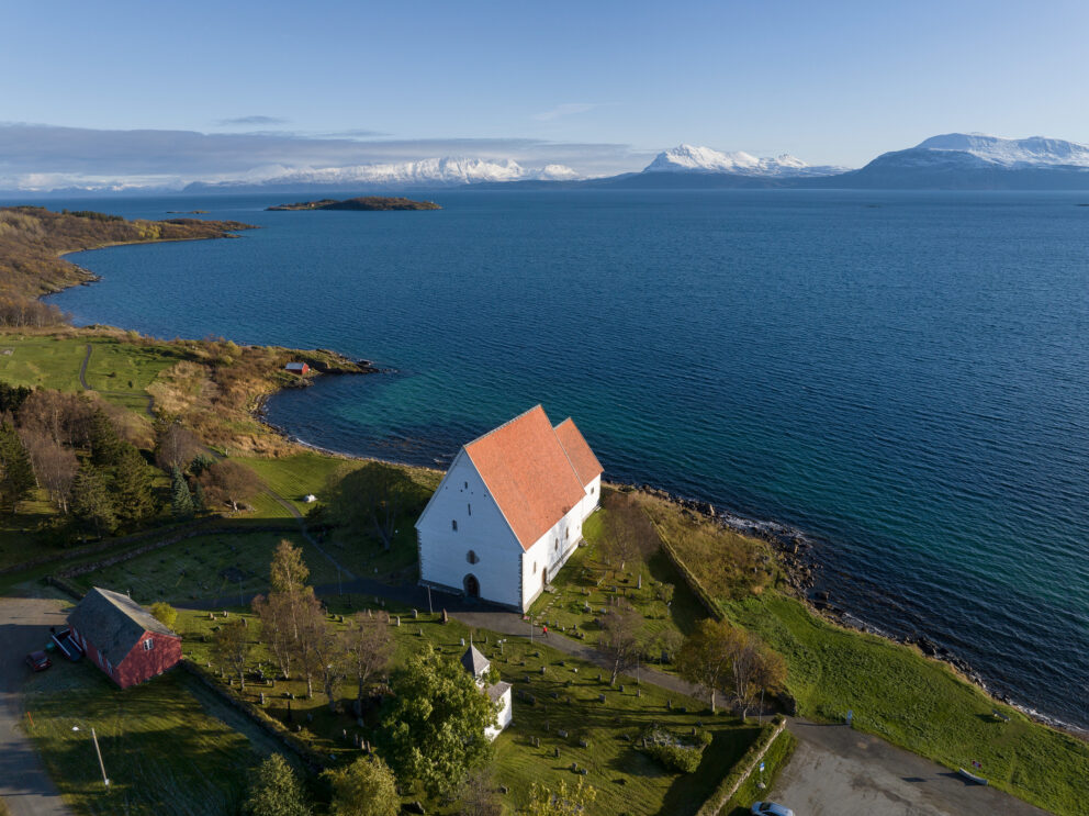 I disse kirkene vil du merke forskjell til jul 5 En hvit steinkirke med rødt tak ligger nær strandkanten, omgitt av grønt gress og trær, med fjell og blått vann i bakgrunnen.