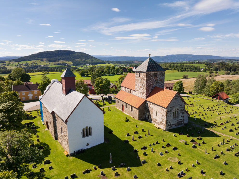 I disse kirkene vil du merke forskjell til jul 7 Flyfoto av to historiske steinkirker med røde tak og en kirkegård i et landlig landskap med åkrer og åser i bakgrunnen.