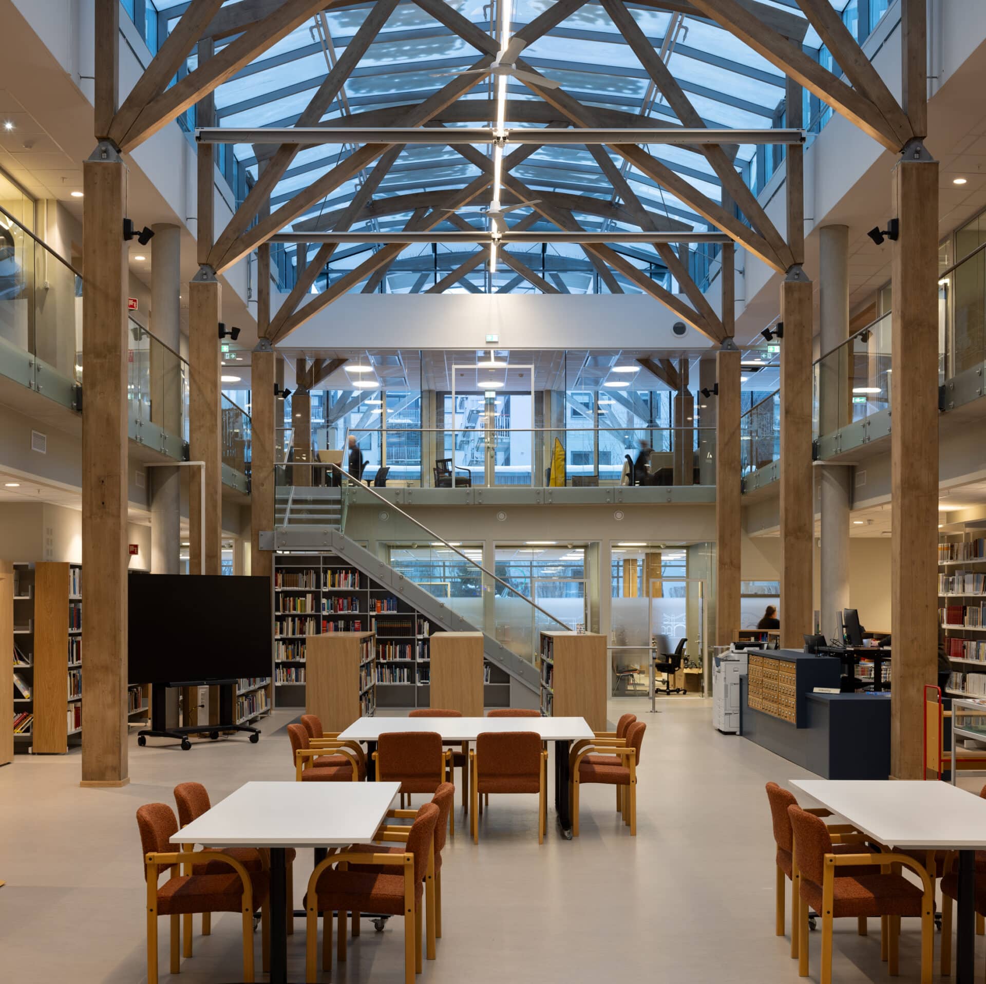 Moderne bibliotekinnredning med synlige trebjelker, glasstak, bokhyller, bord med stoler og en sentral trapp som fører opp til en mesanin.