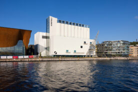 En moderne hvit museumsbygning med navnet Kunstio står ved vannkanten, ved siden av moderne glassbygninger og en oransje buet struktur under en klarblå himmel.