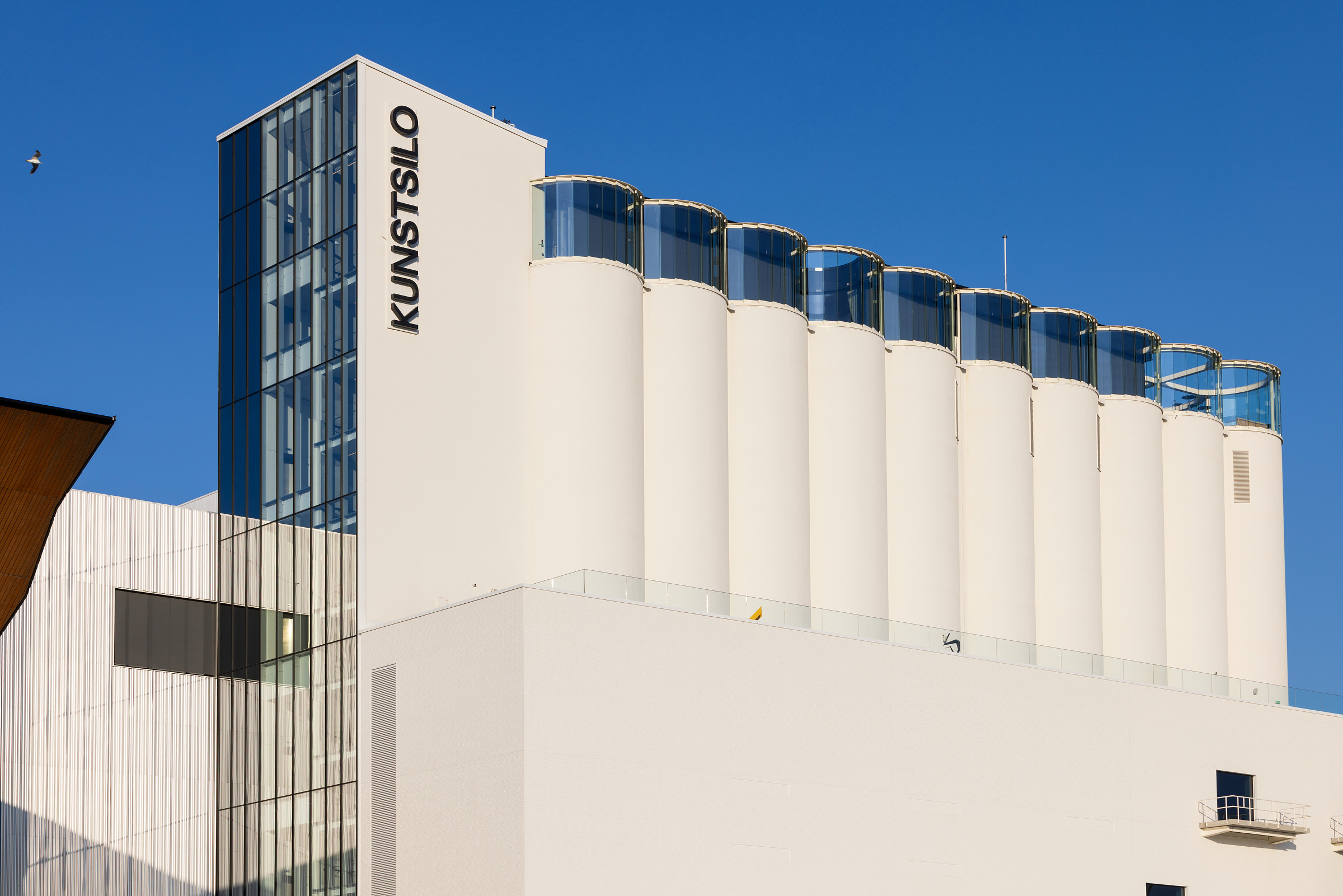 En moderne, hvit bygning merket KUNSTSILO, med en rekke sylinderformede siloer og et hjørne med glassvegger under en klarblå himmel.