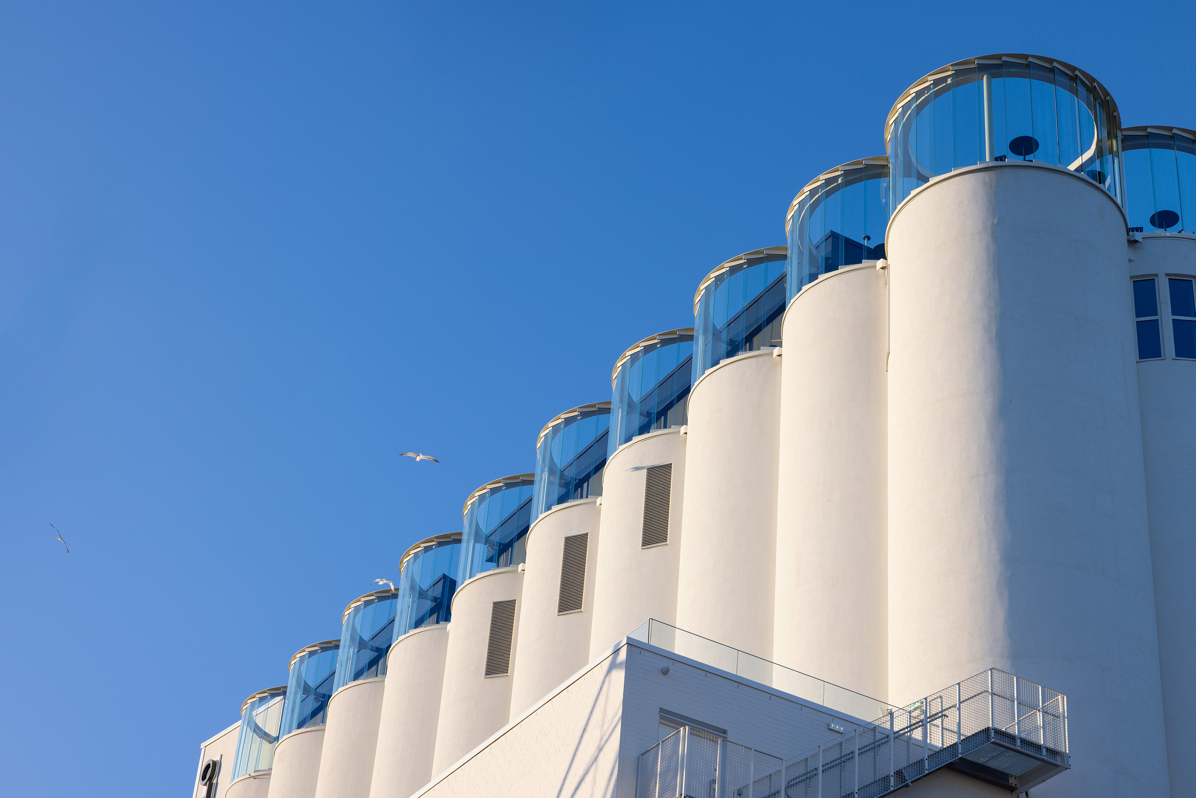 En rekke hvite, sylinderformede siloer med glassoverbygde observasjonsdekk på toppen, mot en klarblå himmel, med noen fugler som flyr i nærheten.