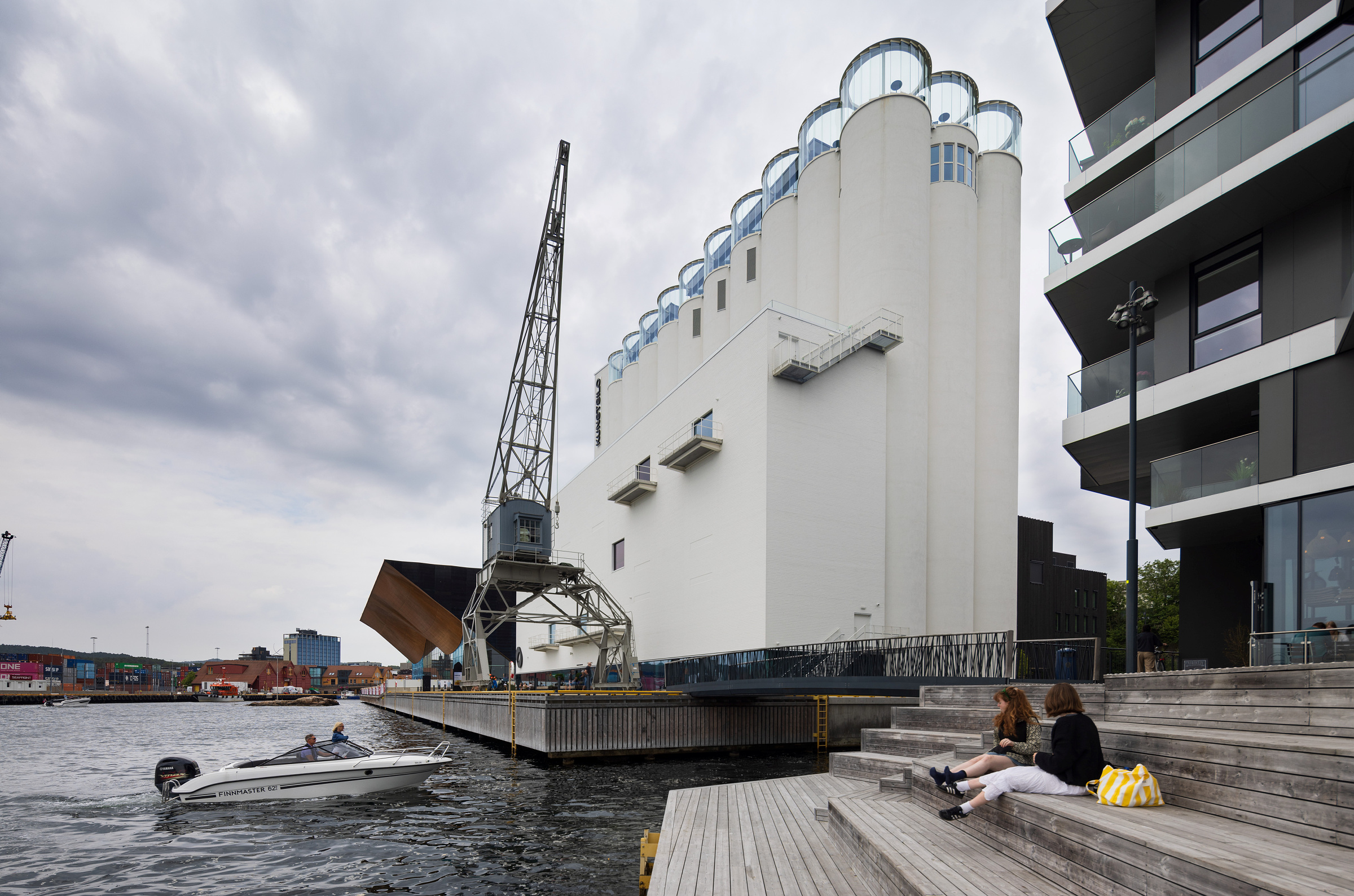 To personer sitter på en trapp ved vannkanten i nærheten av en moderne, hvit bygning i industristil, mens en liten motorbåt passerer forbi og en kran står like ved under en overskyet himmel.