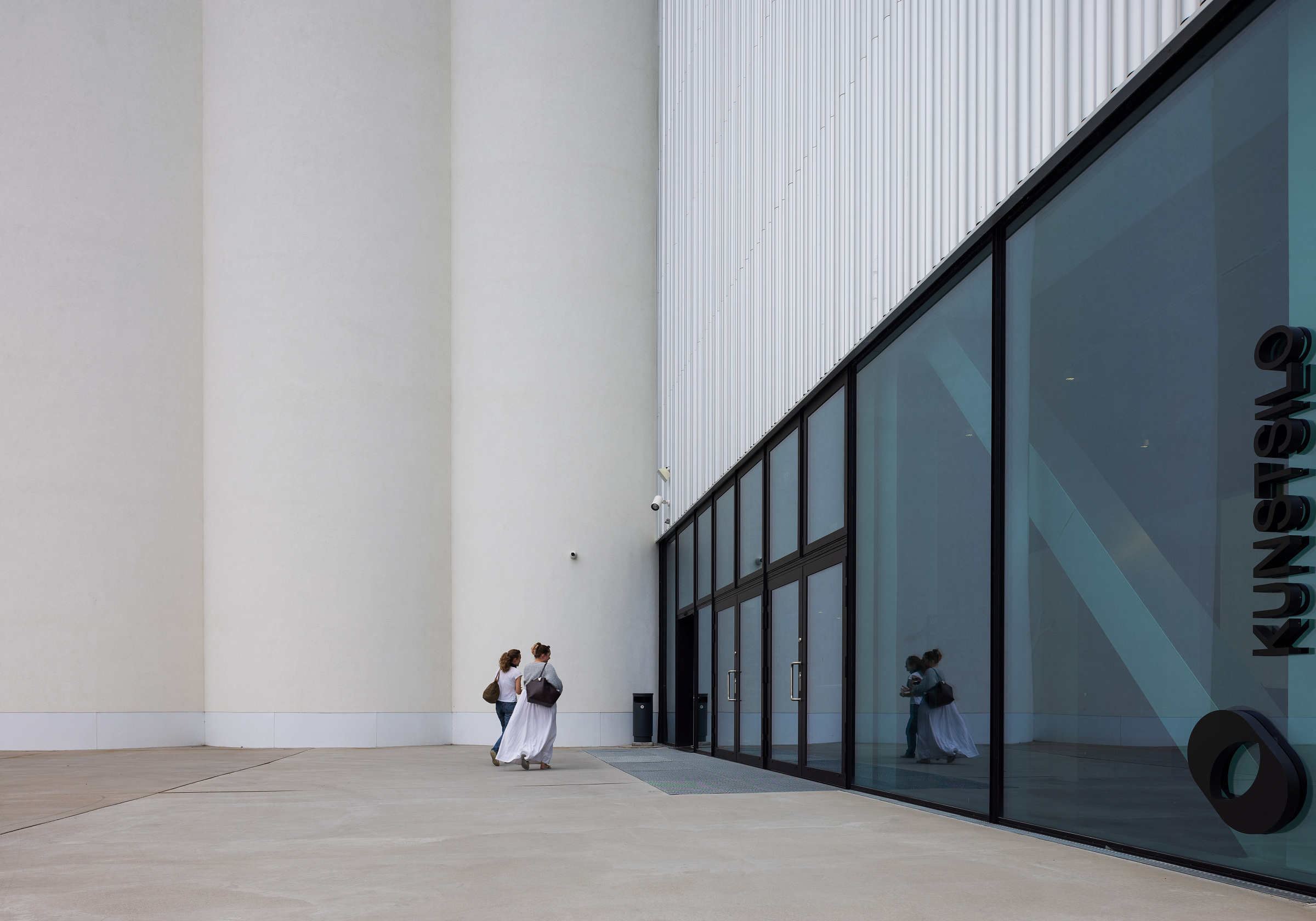 To personer i hvite klær går langs et betongområde utenfor en moderne bygning med store vinduer merket KUNSTMUSEUM.