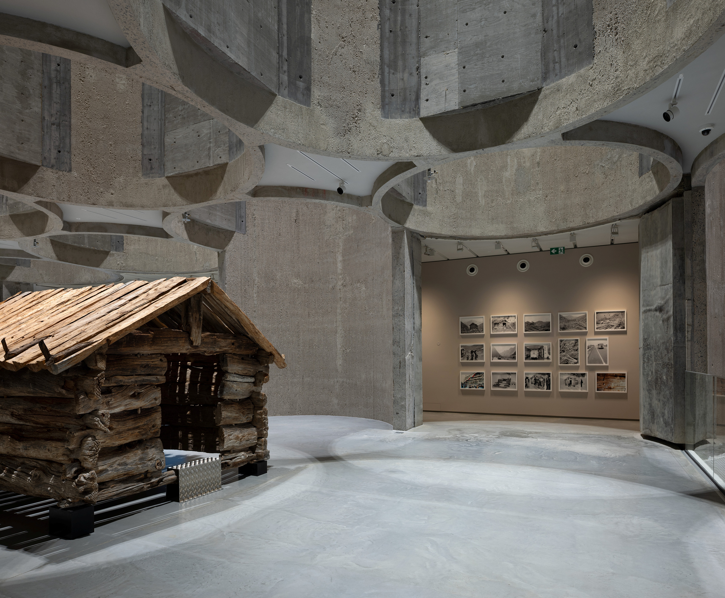 Interiør i et moderne betonggalleri med en liten tømmerhytteskulptur i forgrunnen og innrammede fotografier på den bortre veggen.