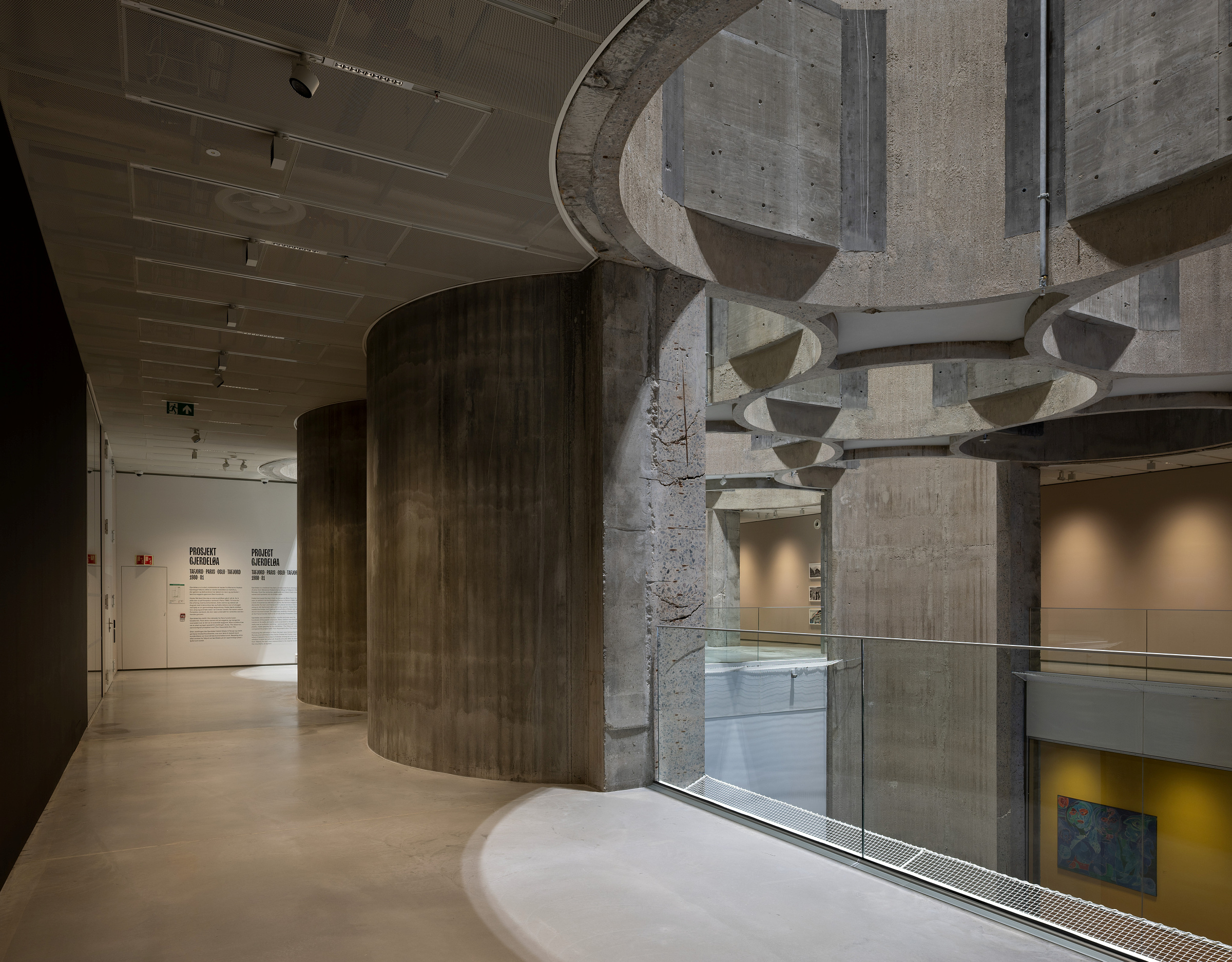 Et moderne interiør med buede betongvegger, store søyler, glassrekkverk og en mesanin med utsikt over et åpent gallerirom under.