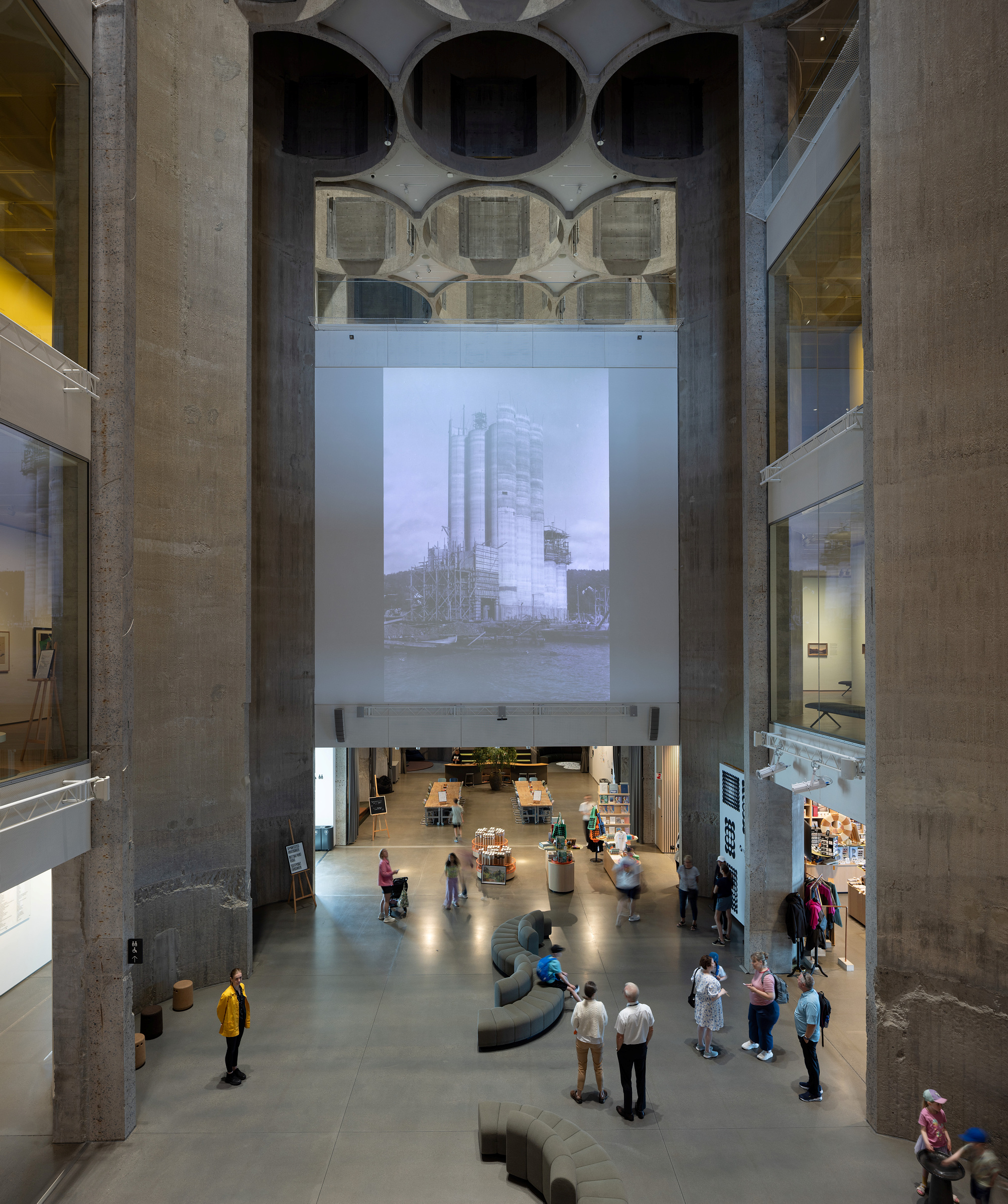 Et romslig atrium i industristil med stor takhøyde, folk som går og sitter, og et stort svart-hvitt fotografi projisert på bakveggen.