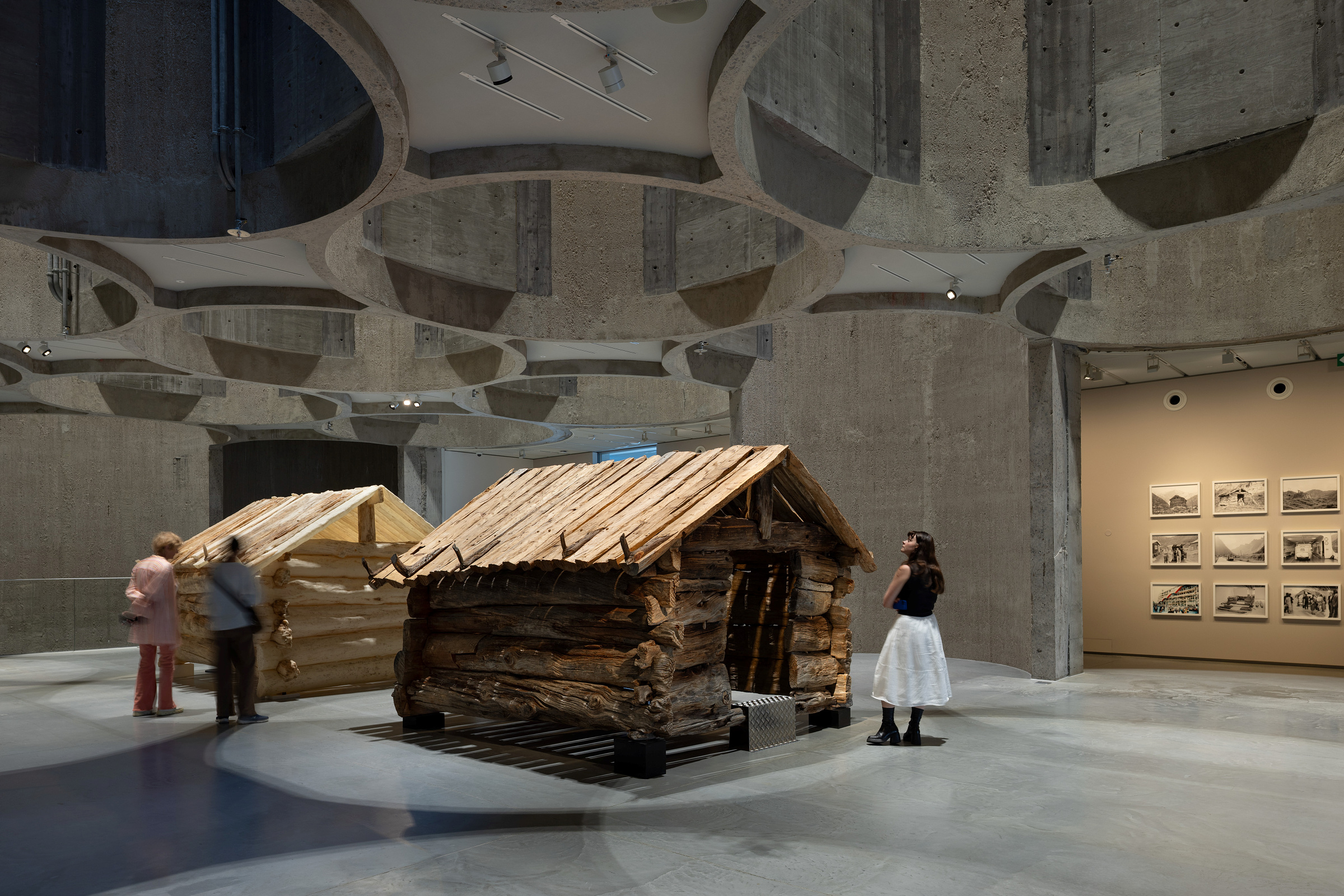 I et moderne gallerirom i betong med sirkulære takutskjæringer ser folk på to hyttekonstruksjoner i tre, mens innrammede fotografier henger på en vegg i bakgrunnen.