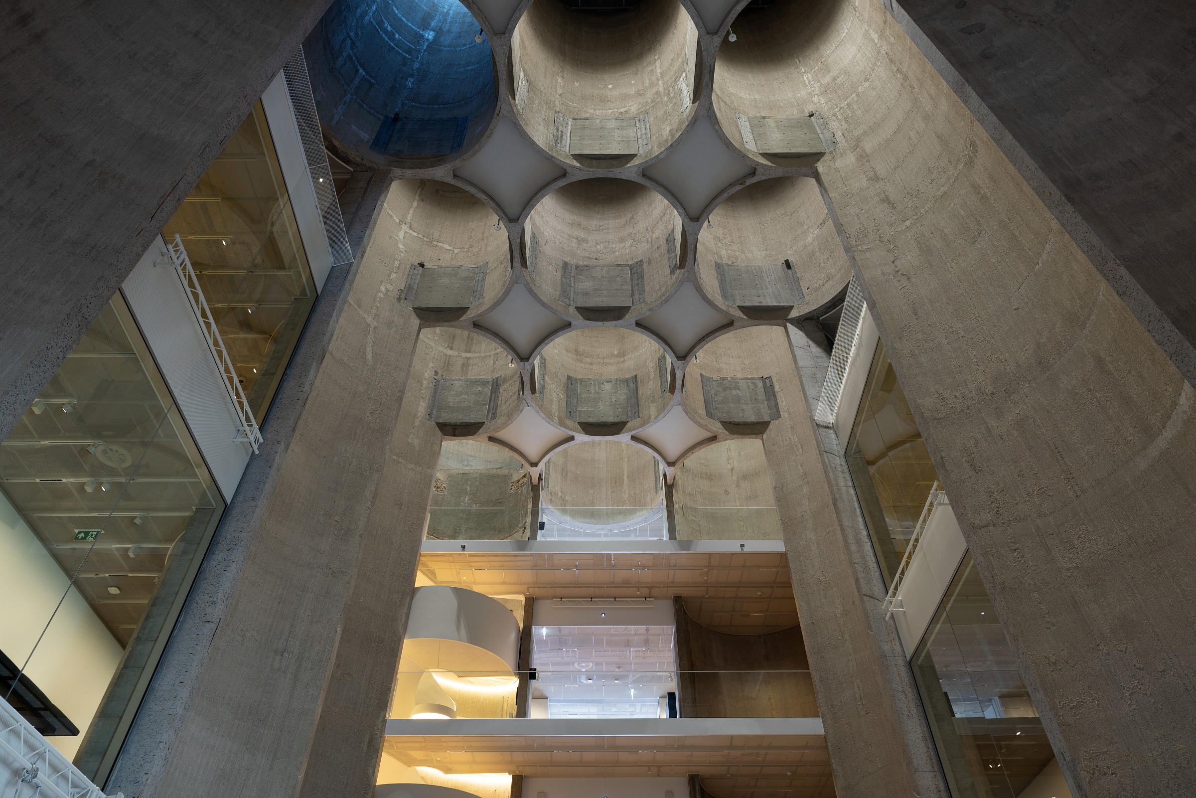 Utsikt opp i en moderne bygning med høye betongvegger, geometriske takutskjæringer, glassbalkonger og naturlig lys som kommer inn ovenfra.