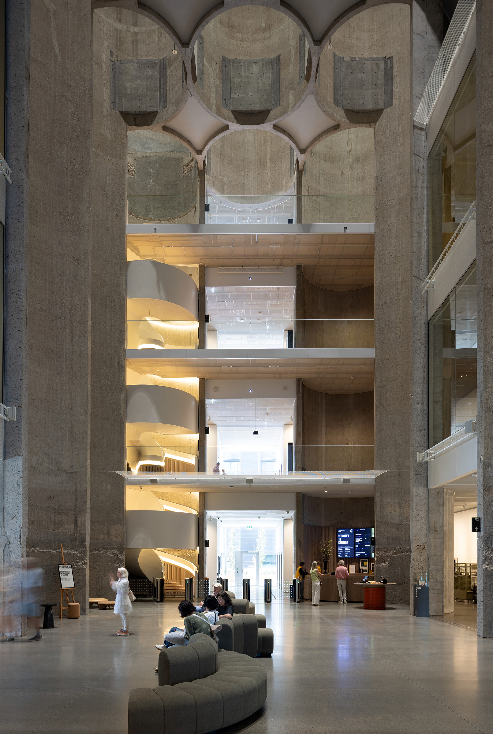 Et romslig, moderne bygningsinteriør med høye betongvegger, flere balkonger, en vindeltrapp og mennesker som sitter eller går i det åpne, sentrale området.