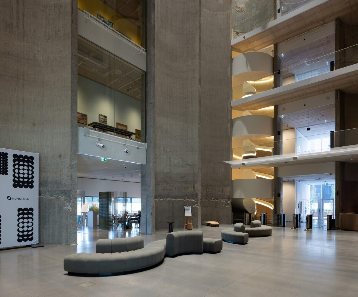 Romslig, moderne lobby med betongsøyler, buede, grå sitteplasser, åpne overetasjer med glassrekkverk og en kafeteria som skimtes i bakgrunnen.