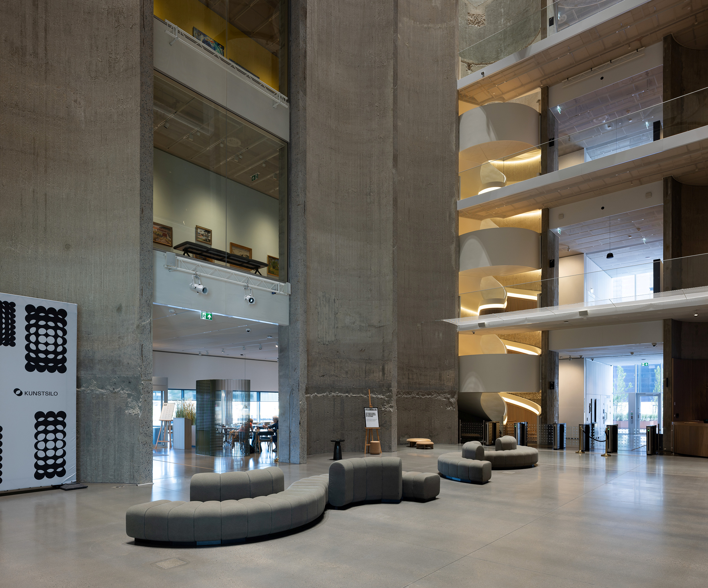 Romslig, moderne lobby med buede, grå sitteplasser, synlige betongsøyler, store vinduer og balkonger i flere nivåer med glassrekkverk og geometrisk belysning.