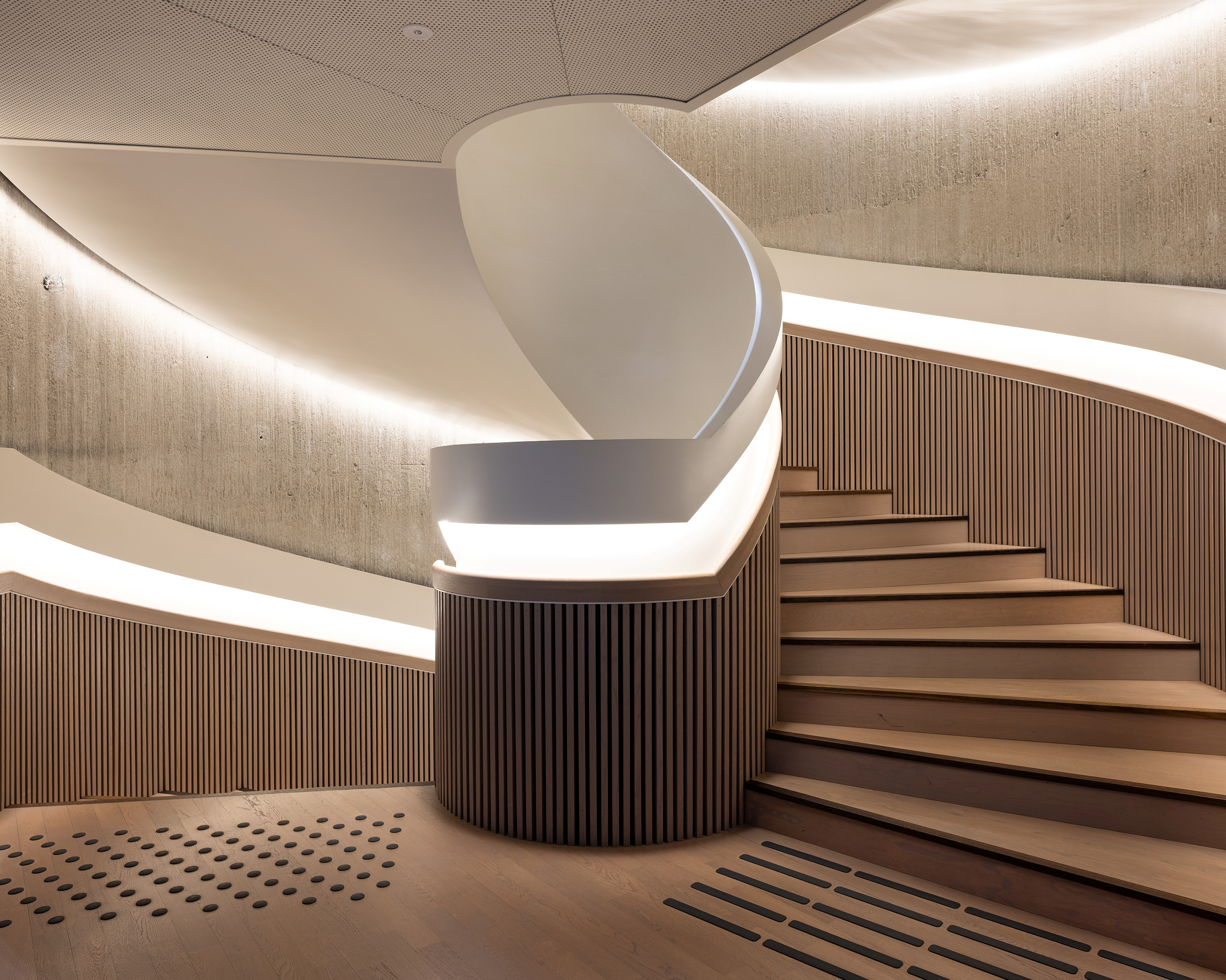 En moderne, buet trapp med tretrinn og riflet trepanel, opplyst av innfelt belysning, mot en betongvegg.