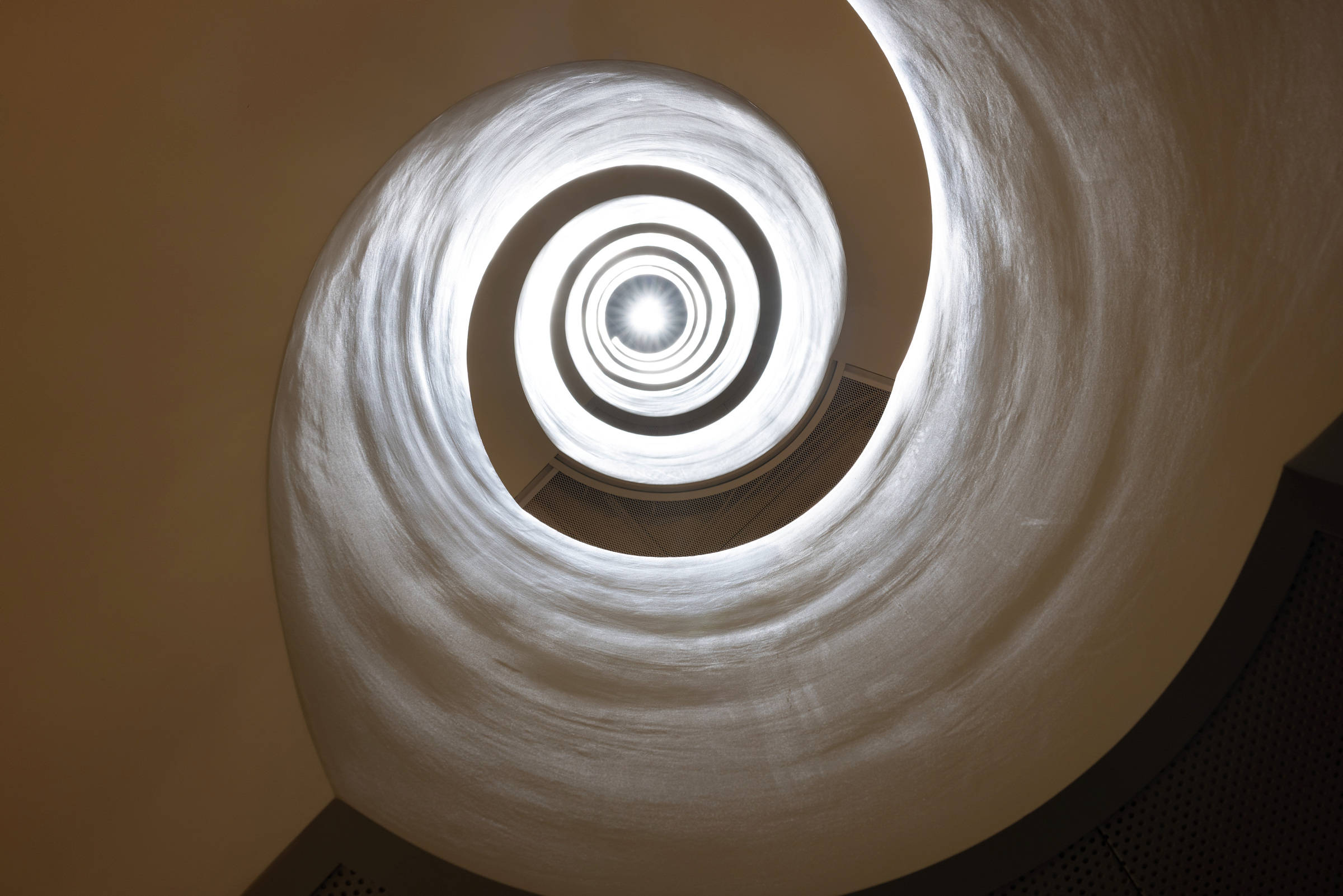 Utsikt opp en vindeltrapp med glatte, hvite vegger, belyst av lys fra midten øverst, noe som skaper en sirkulær, tunnellignende effekt.