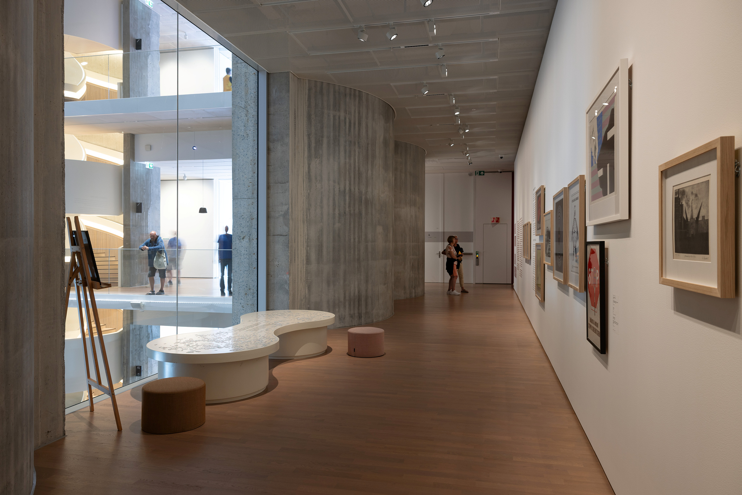 En moderne gallerigang med innrammede kunstverk på veggen, buede sitteplasser og noen få mennesker i bakgrunnen ved store vinduer.