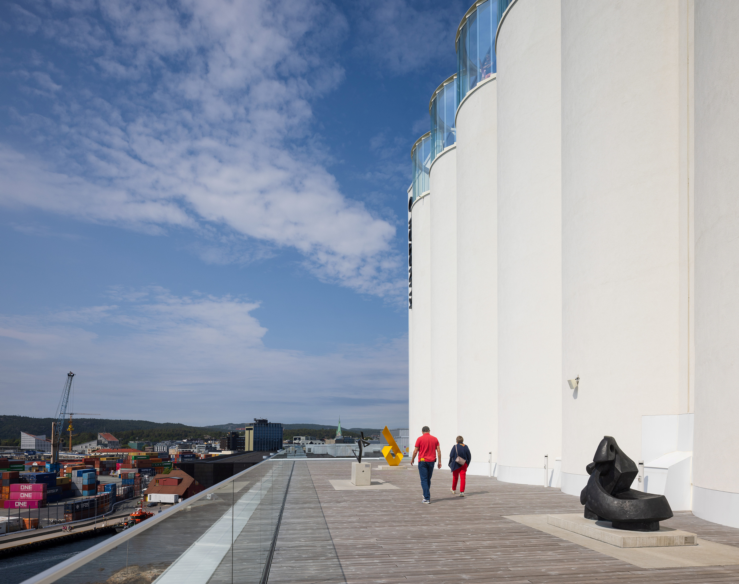 To personer går langs en takterrasse med moderne skulpturer, ved siden av høye, hvite siloer, med utsikt over en havn med containere og kraner under en delvis overskyet himmel.