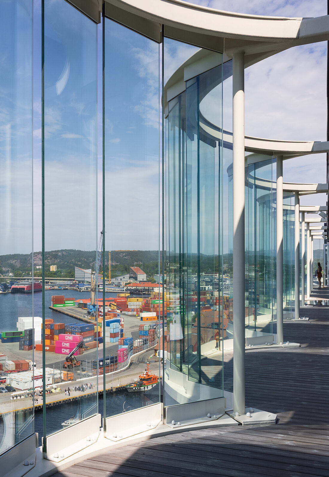 Utsikt over en utskipningshavn med fargerike containere og kraner, sett gjennom buede glassvinduer fra innsiden av en moderne bygning.