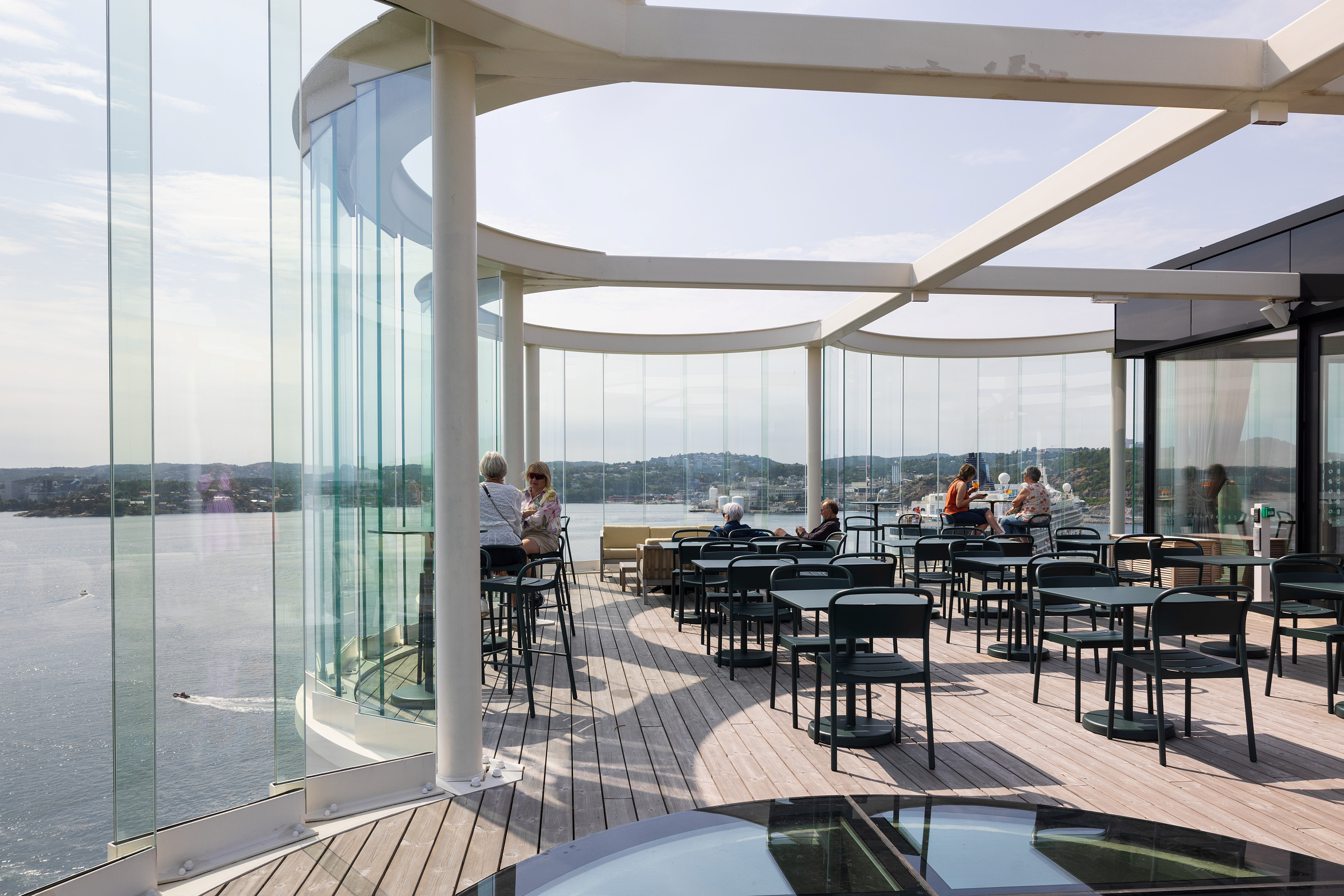Moderne takterrasse med glassvegger, spredte bord og stoler, og folk som sitter og ser ut over et vannlandskap på en solrik dag.