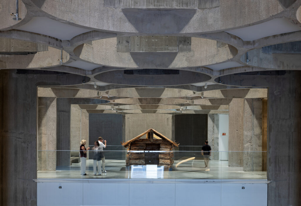 Folk observerer en liten trekonstruksjon inne i et moderne betonginteriør med geometriske takmønstre.