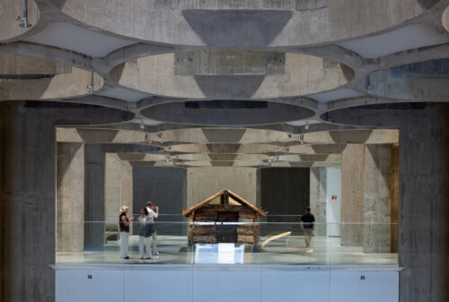 Eksempelsamling 3 Folk observerer en liten trekonstruksjon inne i et moderne betonginteriør med geometriske takmønstre.