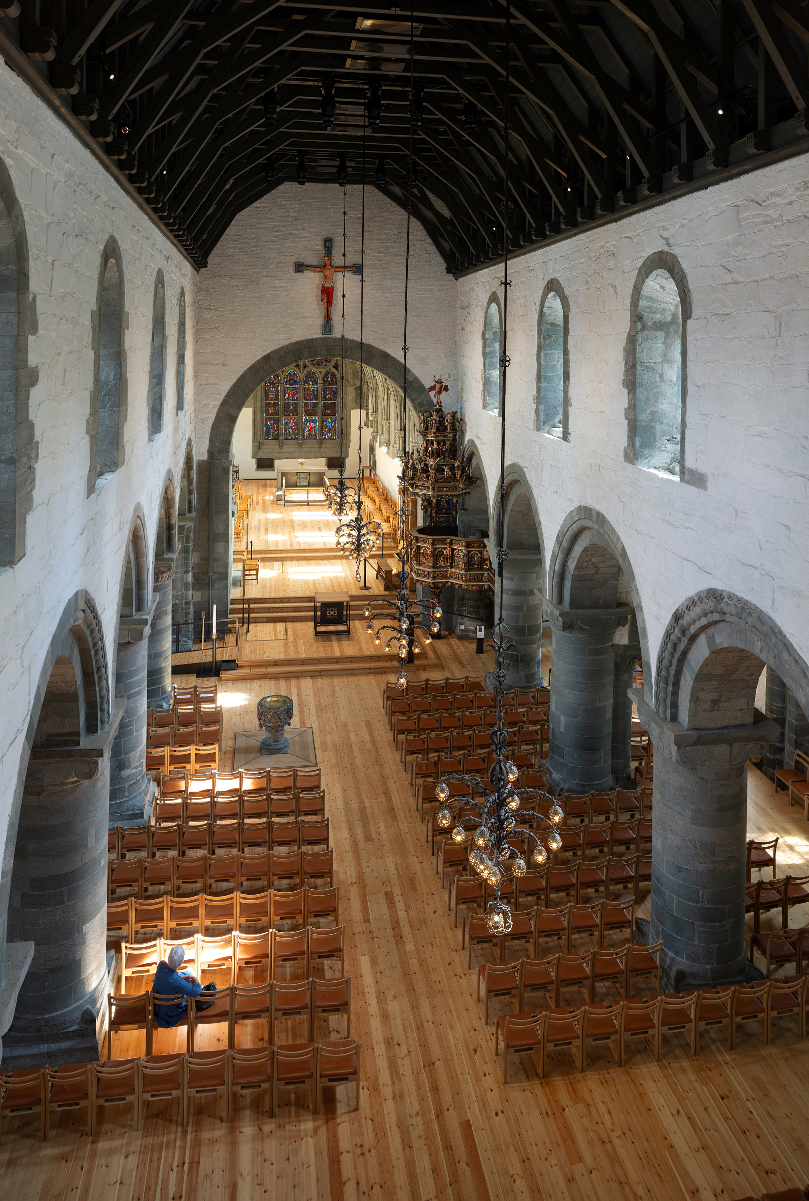 Moderne tilgjengelegheit i mellomalderkyrkje 18 Utsikt over et kirkeinteriør med steinbuer, tretak, glassmalerier, lysekroner og rekker av trestoler, med en person sittende.