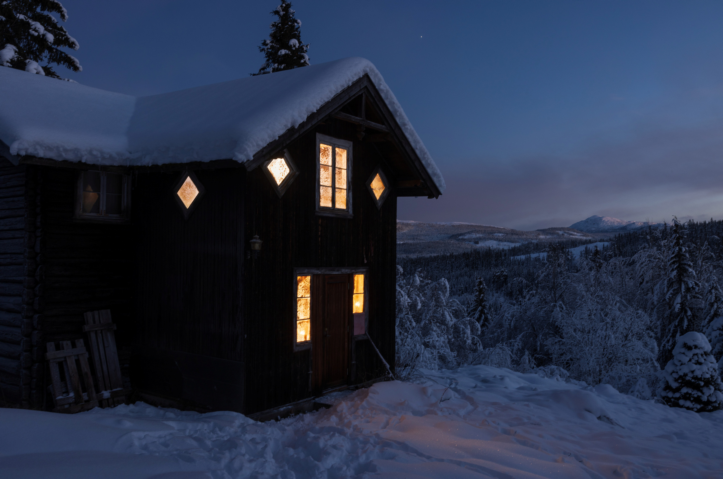 En trehytte med opplyste vinduer står i et snødekt kulturmiljø i skumringen, omgitt av snødekte trær og fjerne åser under en mørknende himmel.