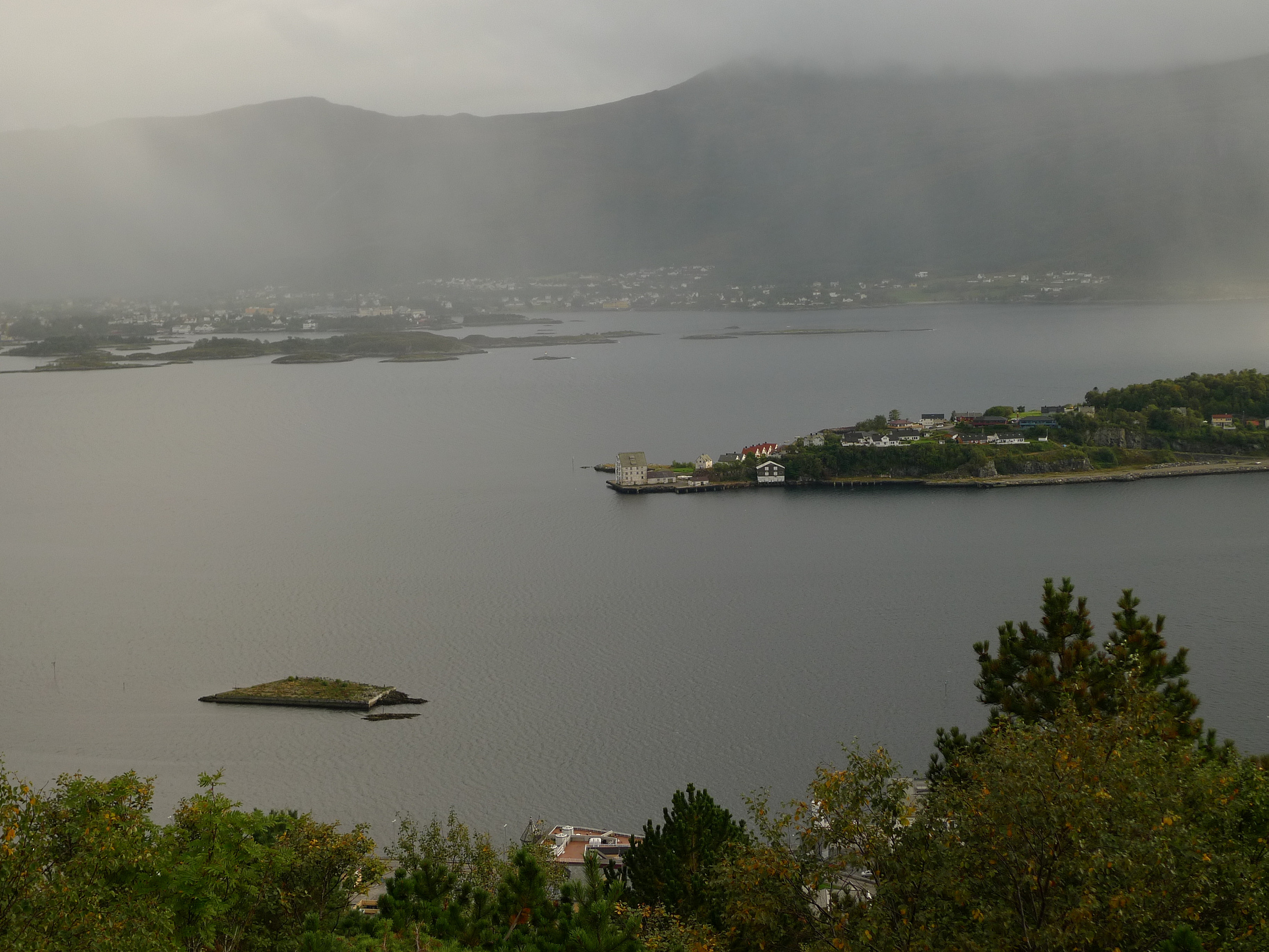 En overskyet utsikt over et rolig vann med små øyer og en by i strandlinjen, omgitt av åser og trær i forgrunnen.